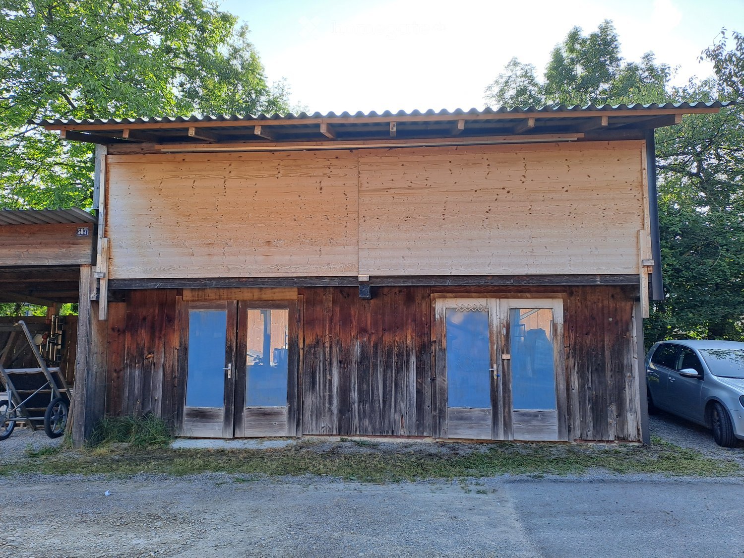 Two-story wooden building with a covered entrance and garage area, surrounded by trees and a parked car.