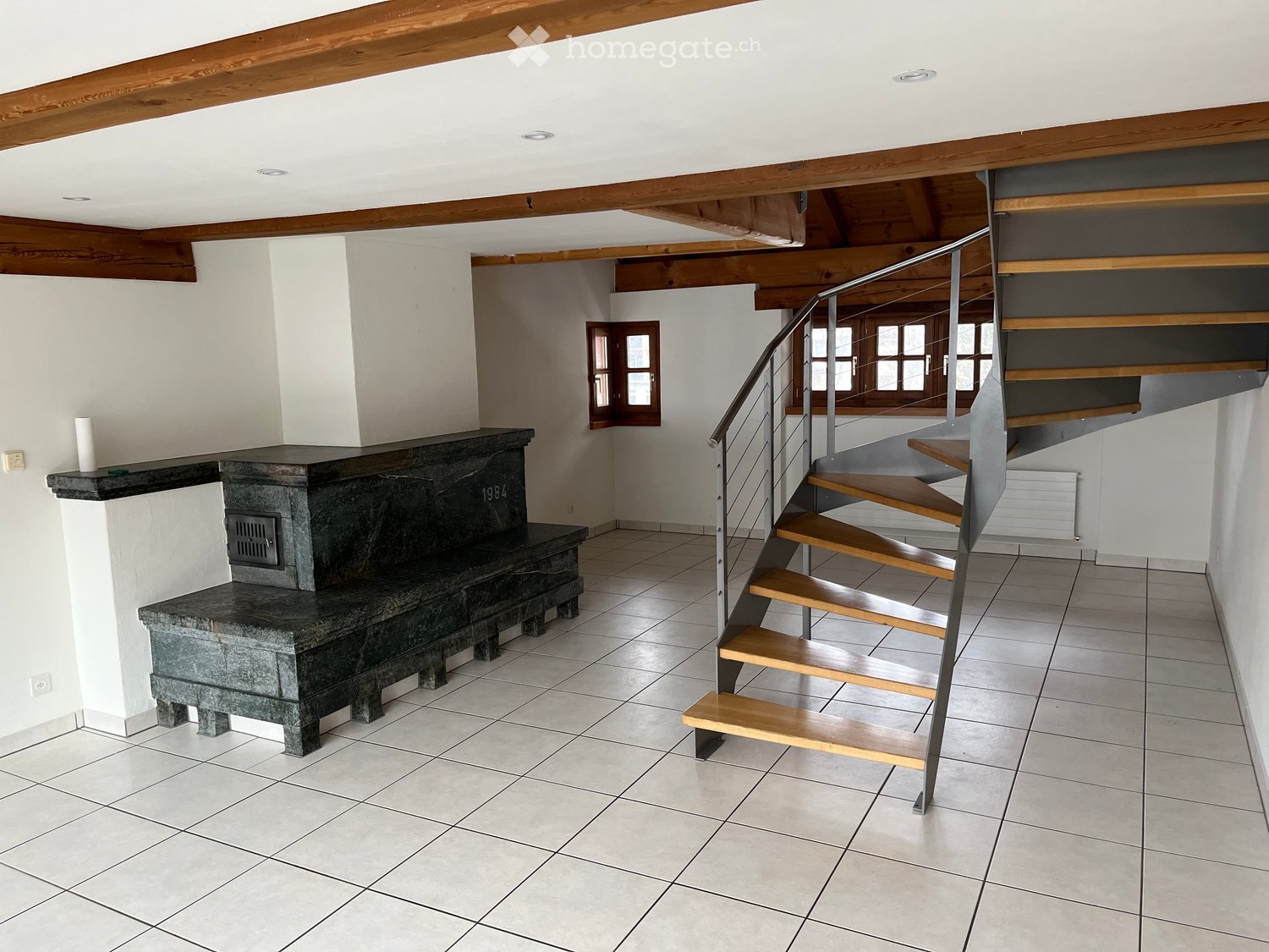 Hallway with exposed wooden beams, tiled floor, staircase with wooden steps and metal railing, and a stone fireplace with a bench.