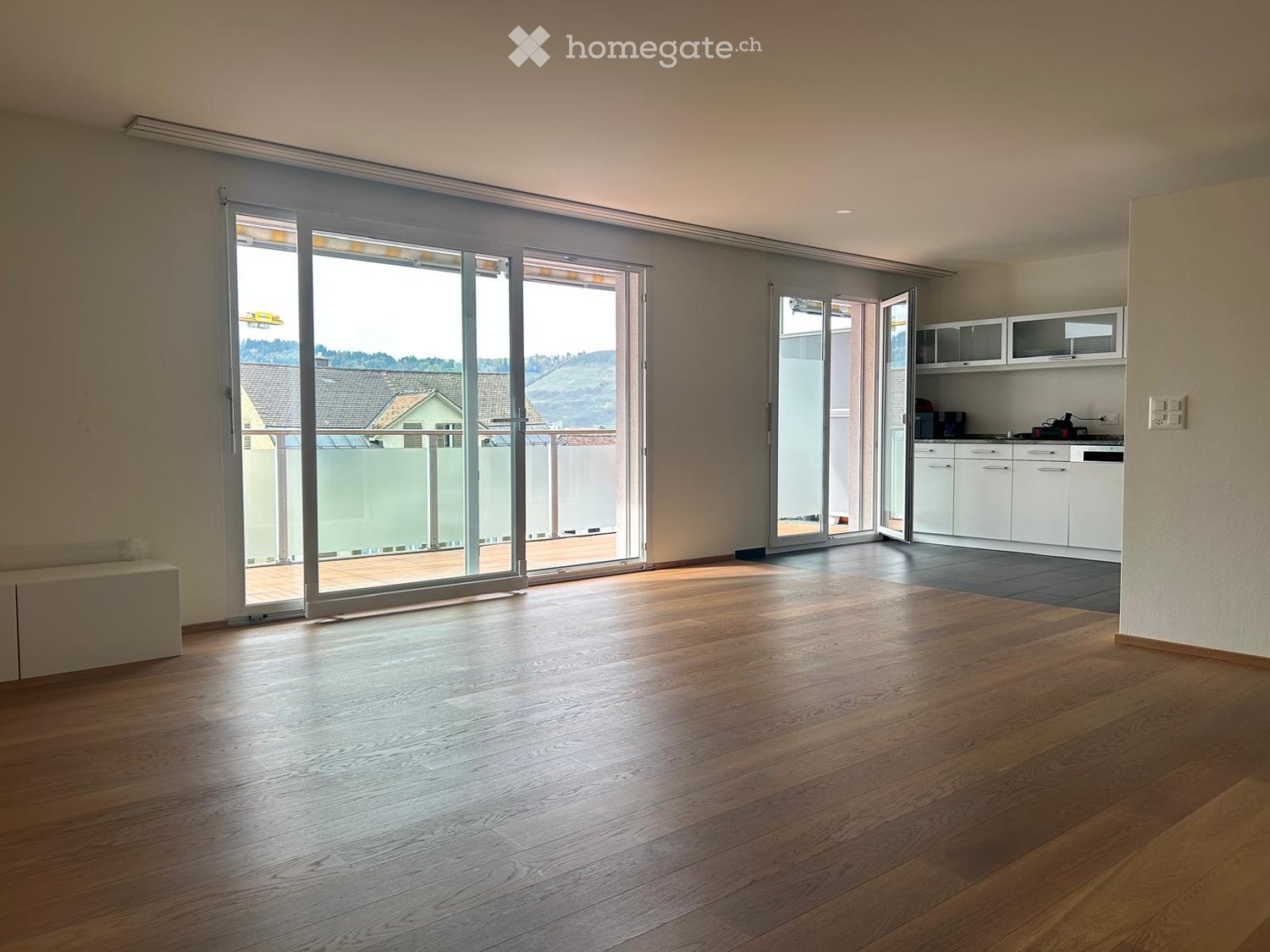 Bright, empty room with wooden flooring, large sliding glass doors leading to a balcony, and a small kitchen area visible in the background.