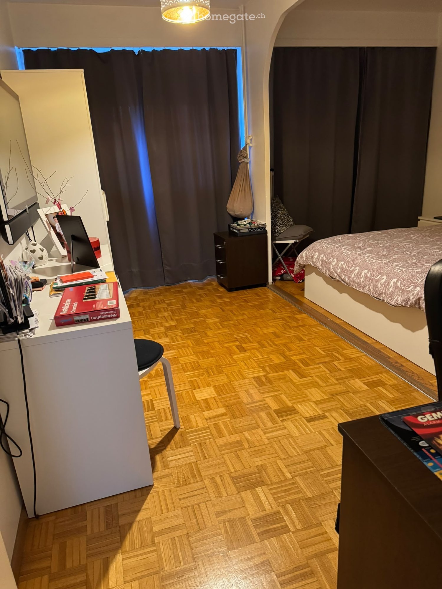 Cozy bedroom with wooden herringbone flooring, a bed with patterned bedding, a desk with a laptop and various items, a nightstand, and a curtained window.