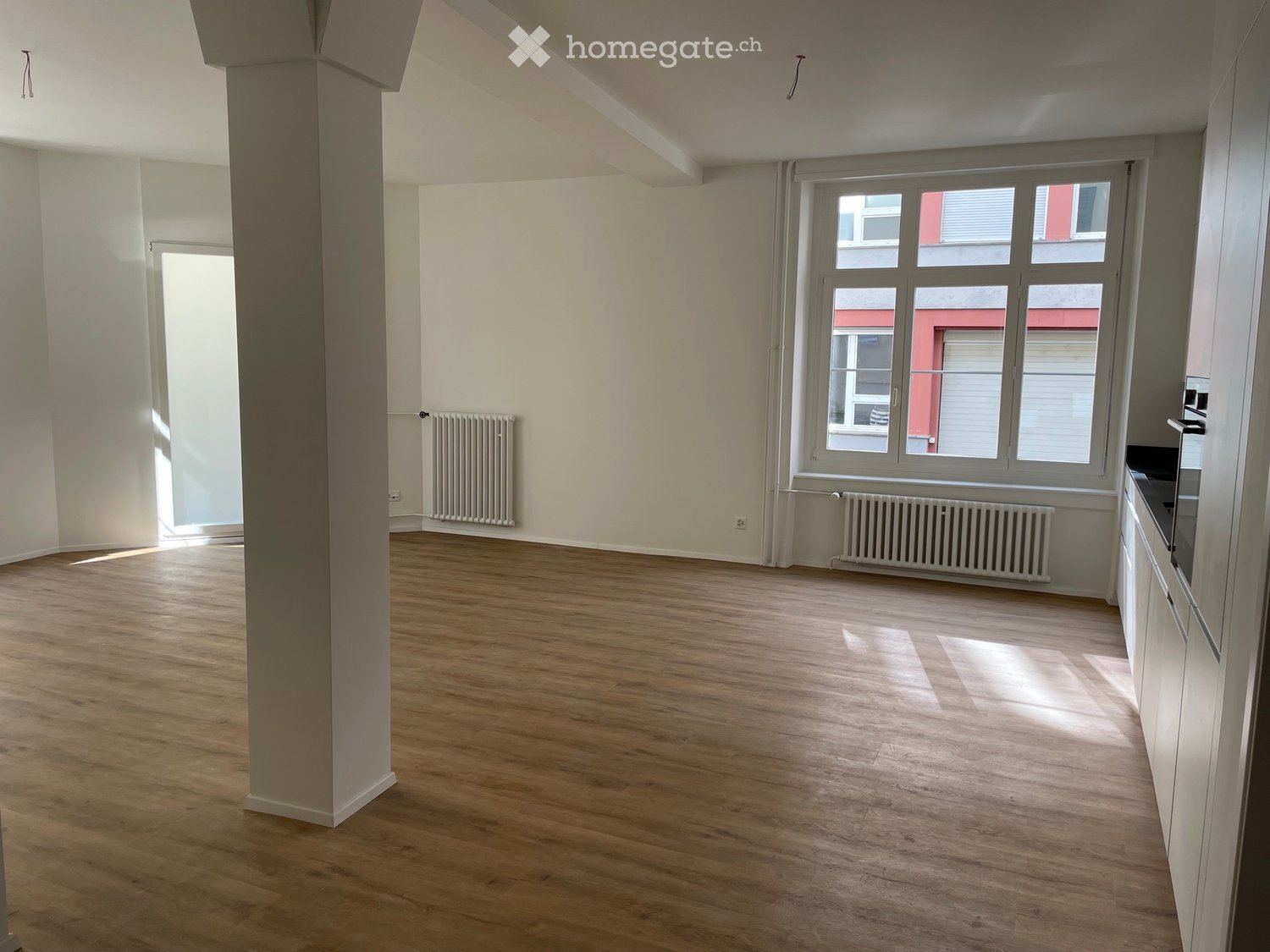 Empty room with wooden flooring, white walls, and large windows allowing natural light. Features include a central column, radiators, and a small kitchen area with cabinets.
