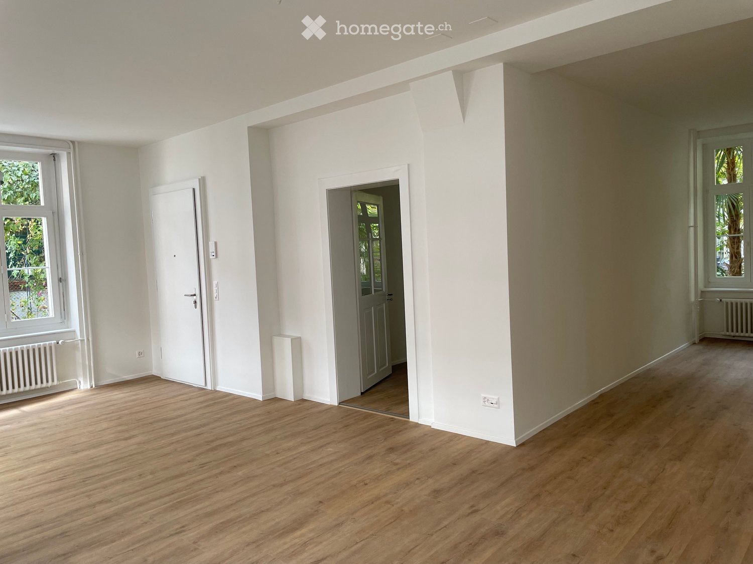 Empty room with light wooden flooring, white walls, and two doors leading to other spaces. Windows on both sides allow natural light to enter.