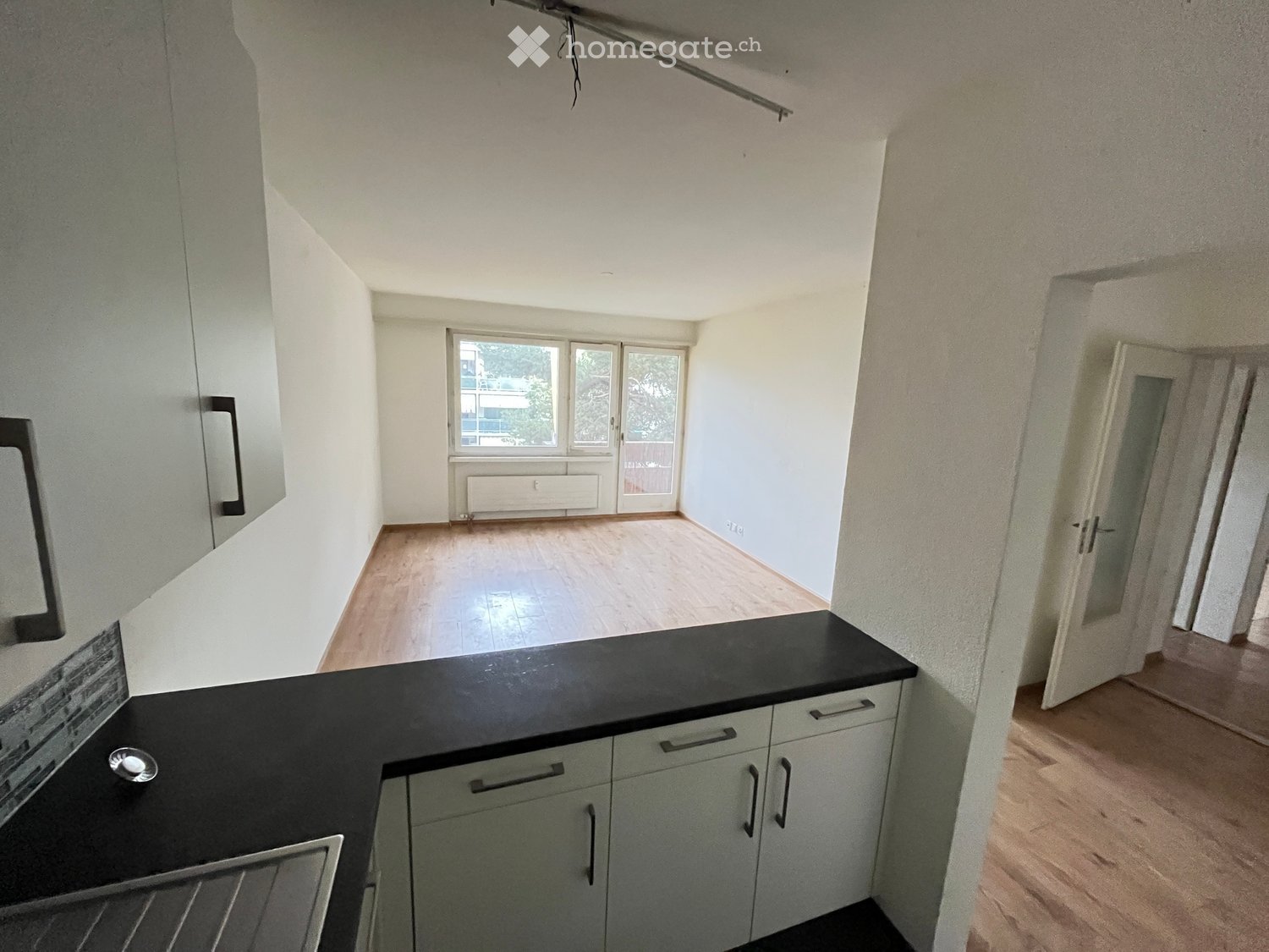 A modern, minimalist kitchen with light grey cabinets, a black countertop, and a wooden floor. The space is open and bright, with large windows and a door leading to another room.