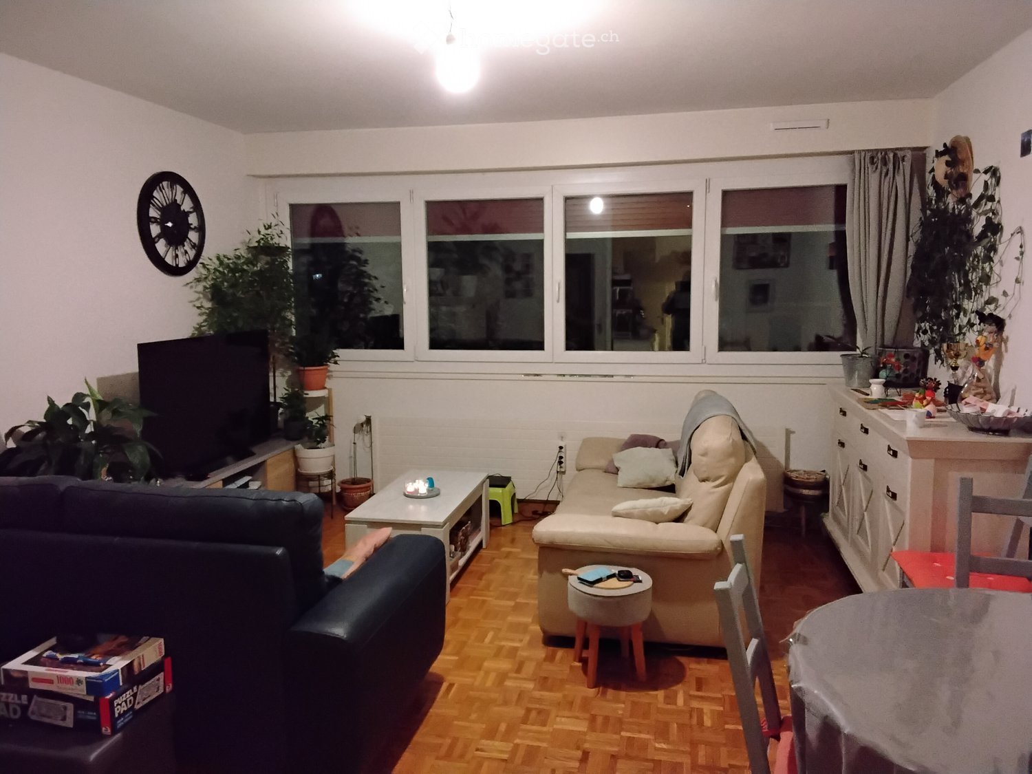 Living room with a beige sofa, coffee table, small stool, and a dark blue armchair. There is a television on a stand, a clock on the wall, and several potted plants near the windows.