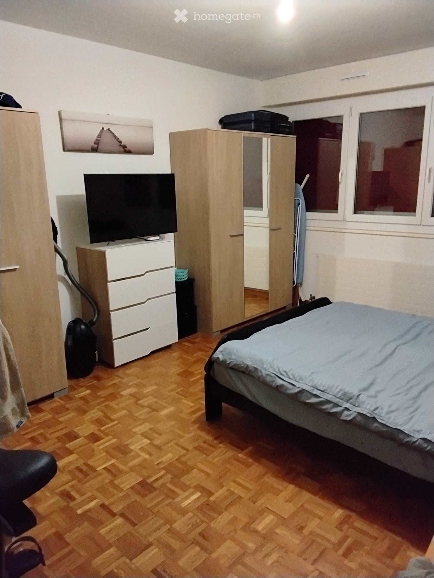 A furnished bedroom with a wooden floor, a bed with blue bedding, a wardrobe, a TV on a stand, and a chair.