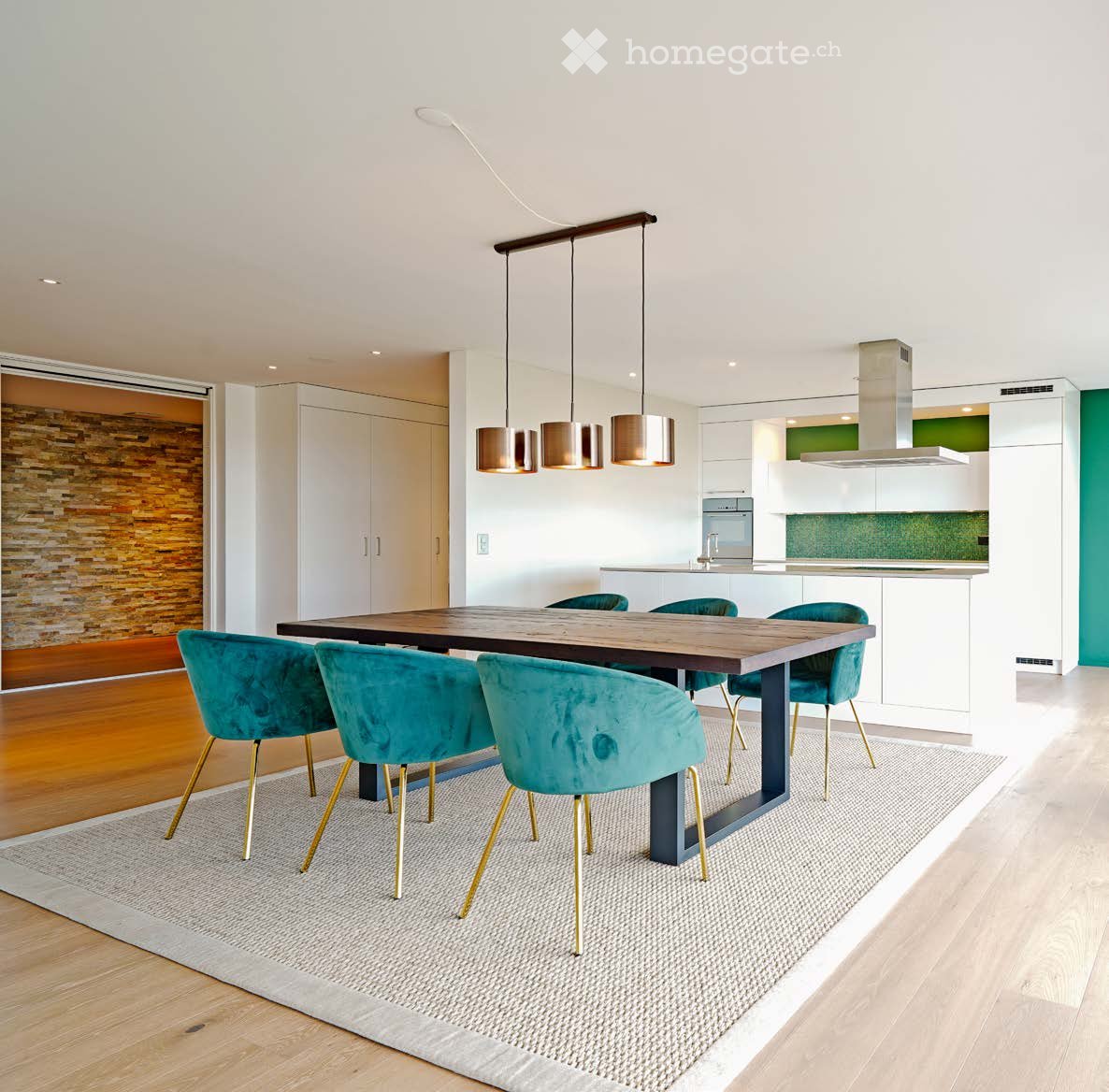 Modern dining area with a wooden dining table, teal upholstered chairs, pendant lights, and an open kitchen with white cabinets and stainless steel appliances.