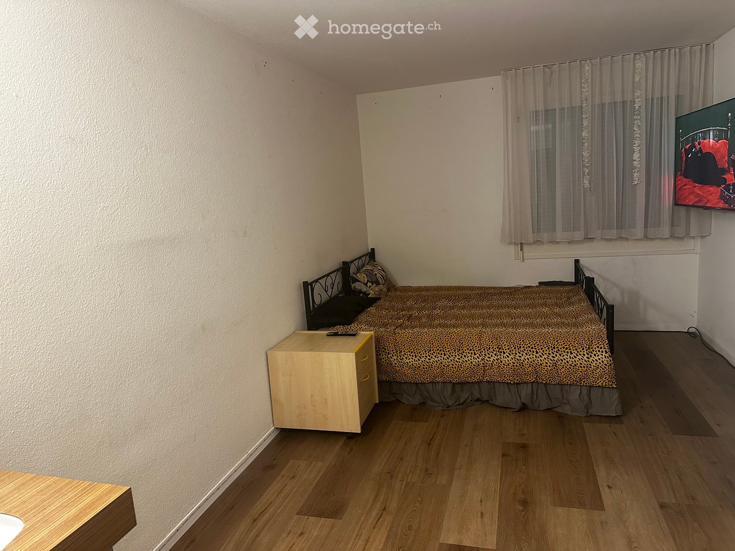 Cozy bedroom with wooden flooring, a single bed with leopard print bedding, a nightstand, and a television mounted on the wall.