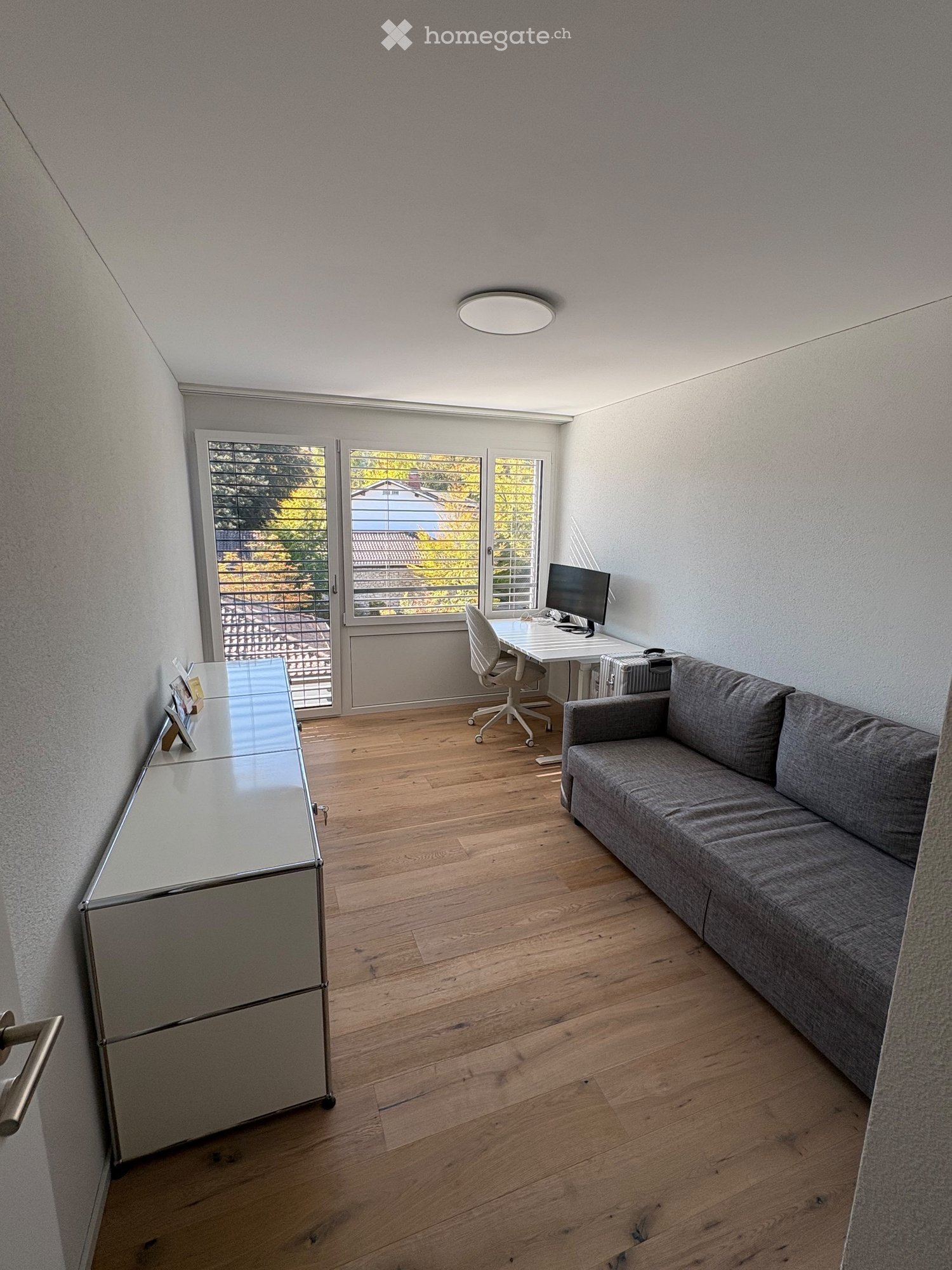 A modern, well-lit room with wooden flooring, a grey sofa, a desk with a computer, and a glass table with a mirror.