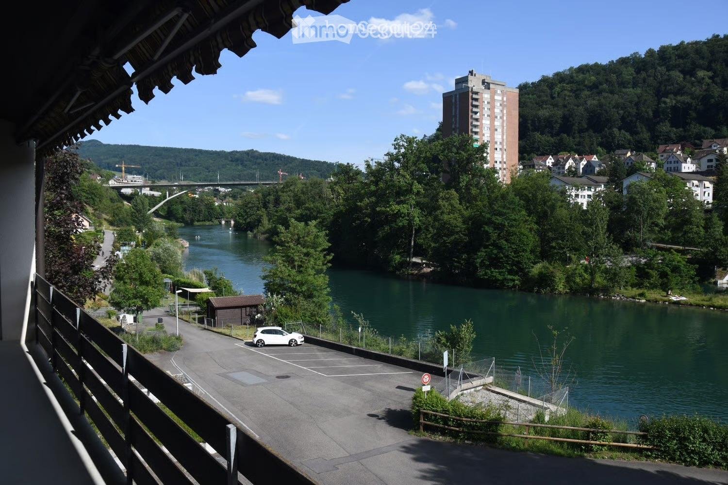 Balcony overlooking a river with a parking area, a small shed, and a distant cityscape with buildings and a bridge.