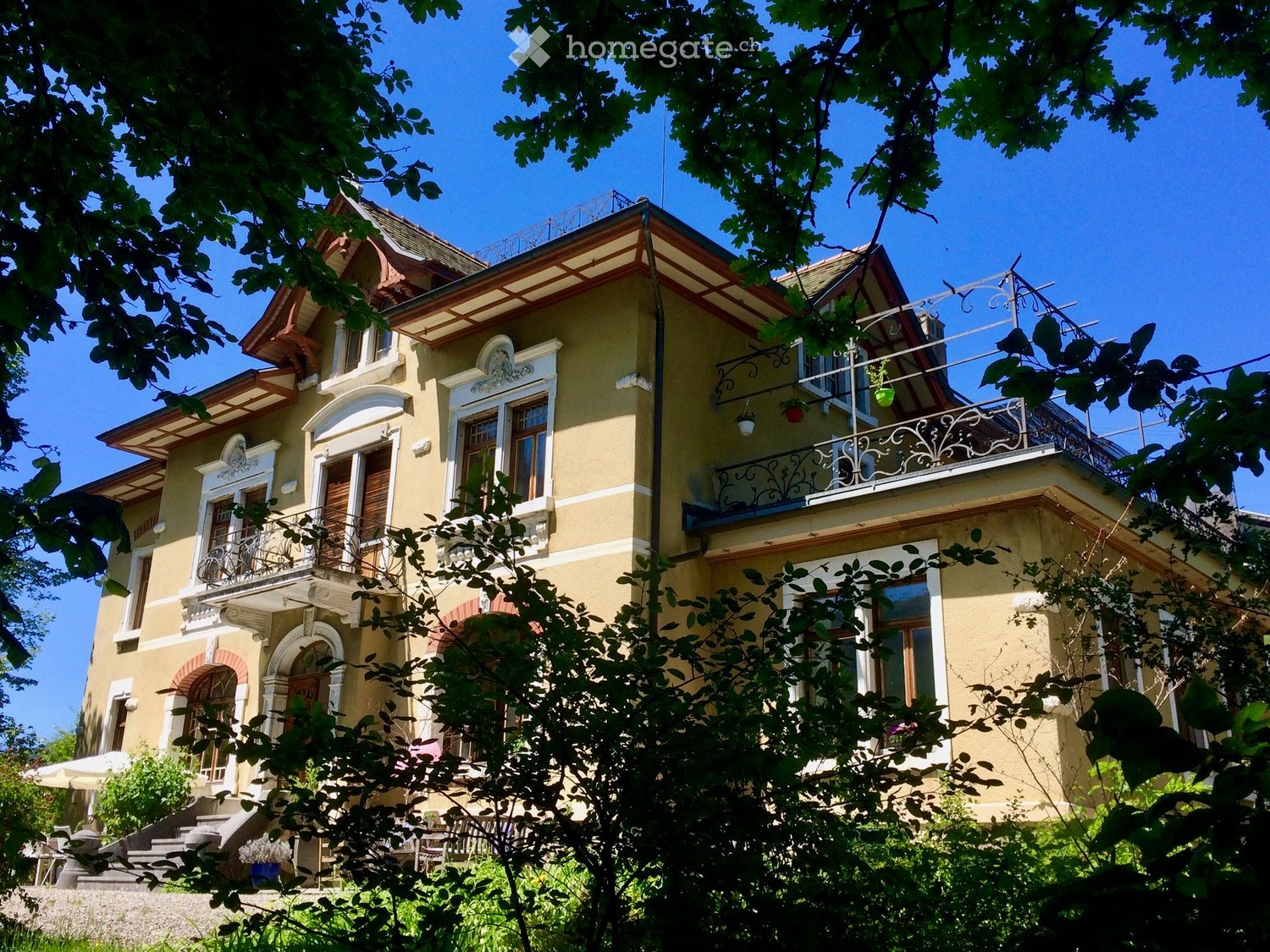 A two-story yellow house with white trim, multiple balconies, ornate railings, and decorative architectural details.