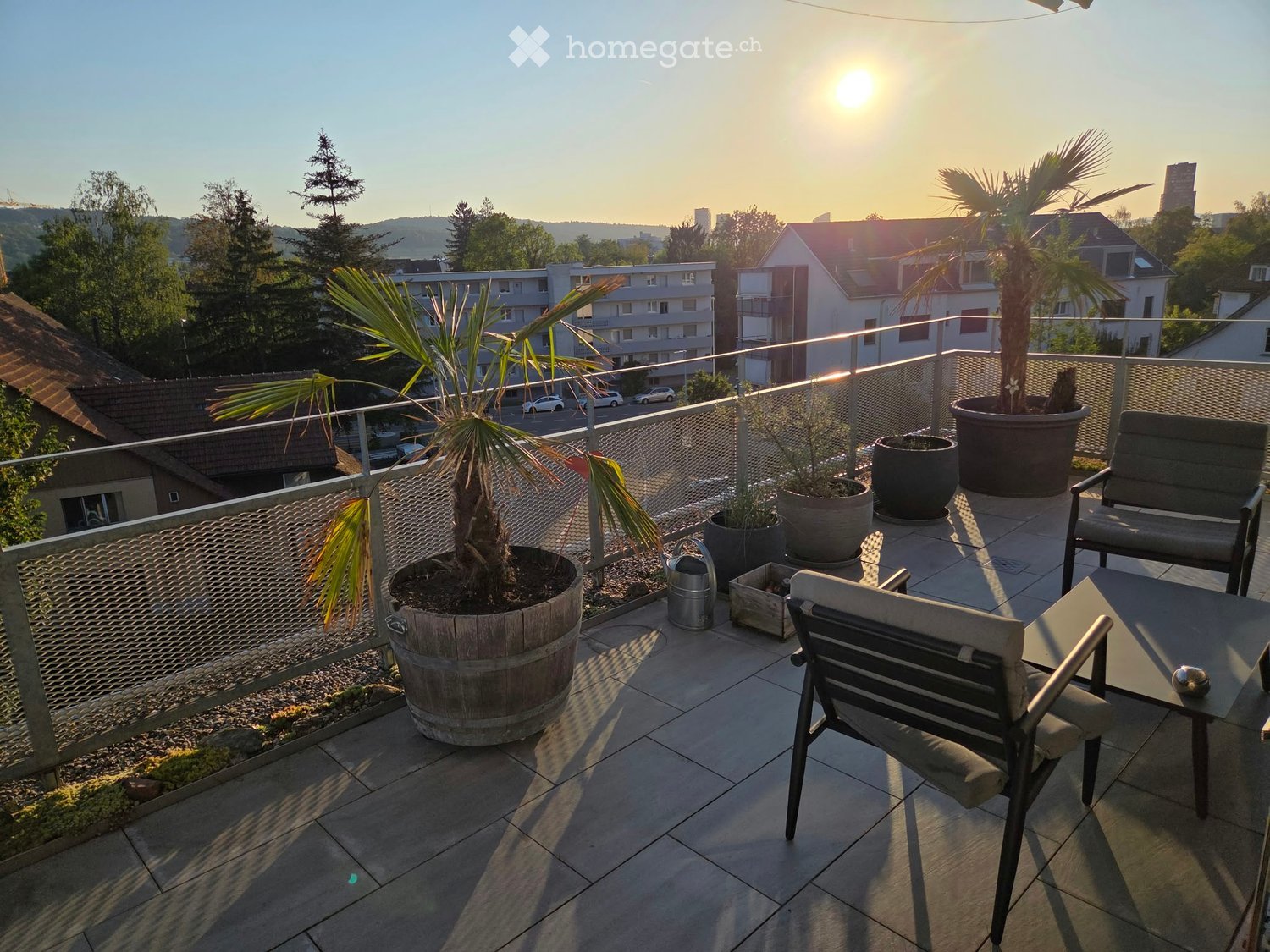 Balcony with tiled floor, metal railing, potted plants, and outdoor furniture including chairs and a table.