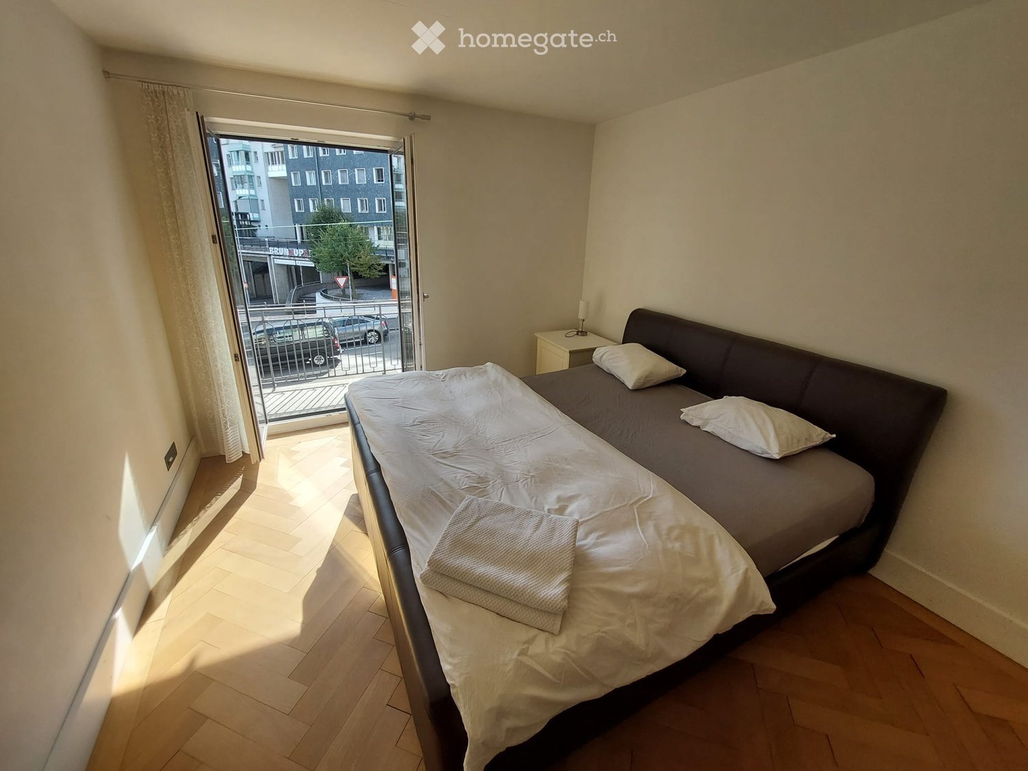 Cozy bedroom with a double bed, nightstand, and lamp. The room features wooden flooring and a large window with a view of the street.