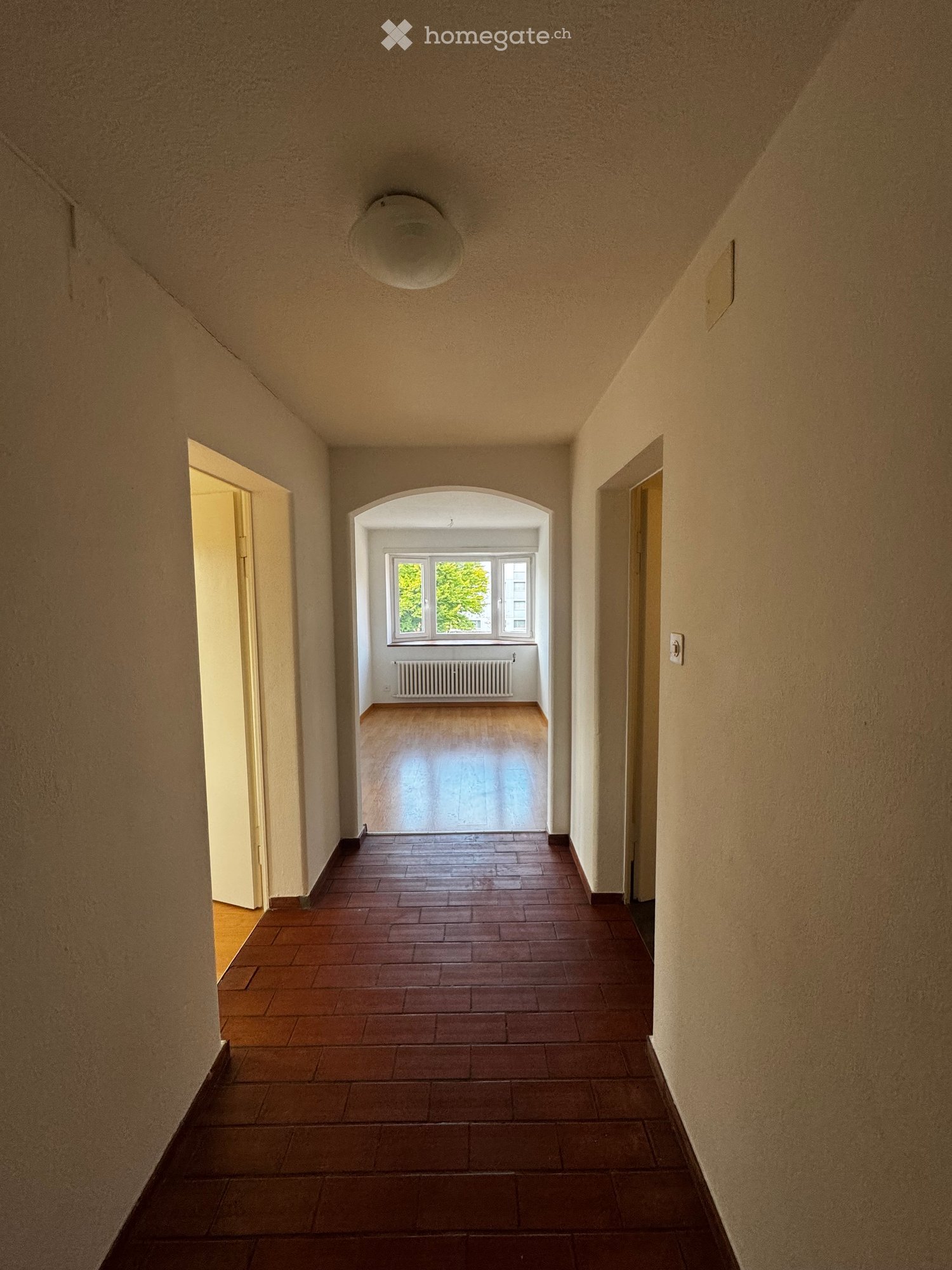 Empty hallway with tiled floor, beige walls, and a ceiling light. An open doorway leads to a room with wooden flooring, a radiator, and a large window showing greenery outside.