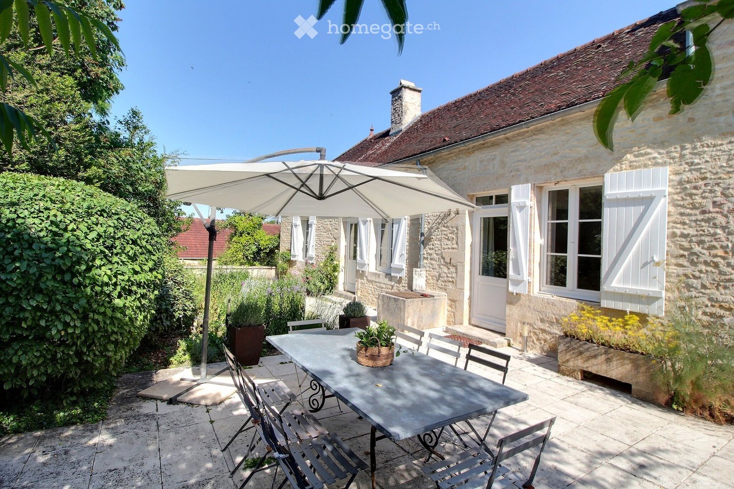 Outdoor patio area with a large table, chairs, umbrella, and potted plants, adjacent to a stone house with white shutters and a chimney.