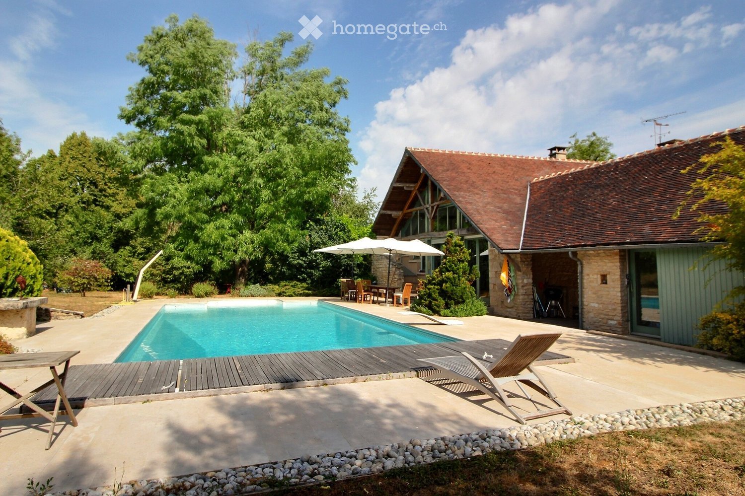 Outdoor area with a swimming pool, wooden deck, lounge chairs, umbrella, and dining set under trees.