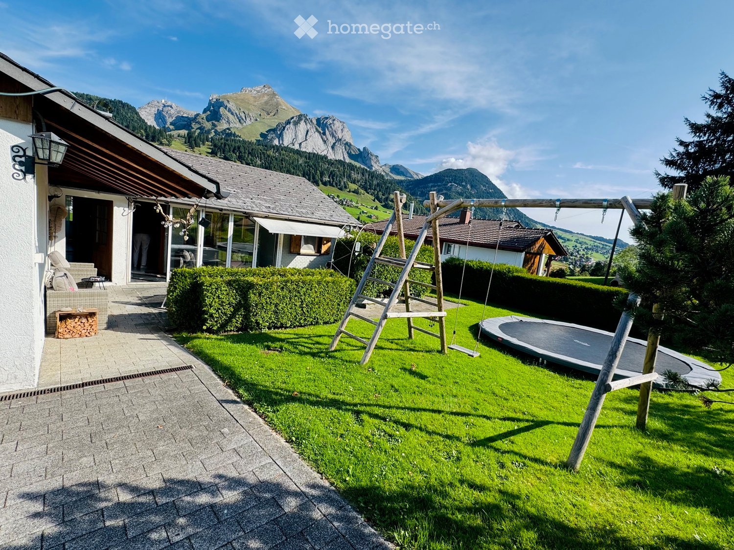 Lush green garden with a swing set, trampoline, neatly trimmed hedges, and a view of snow-capped mountains in the background.