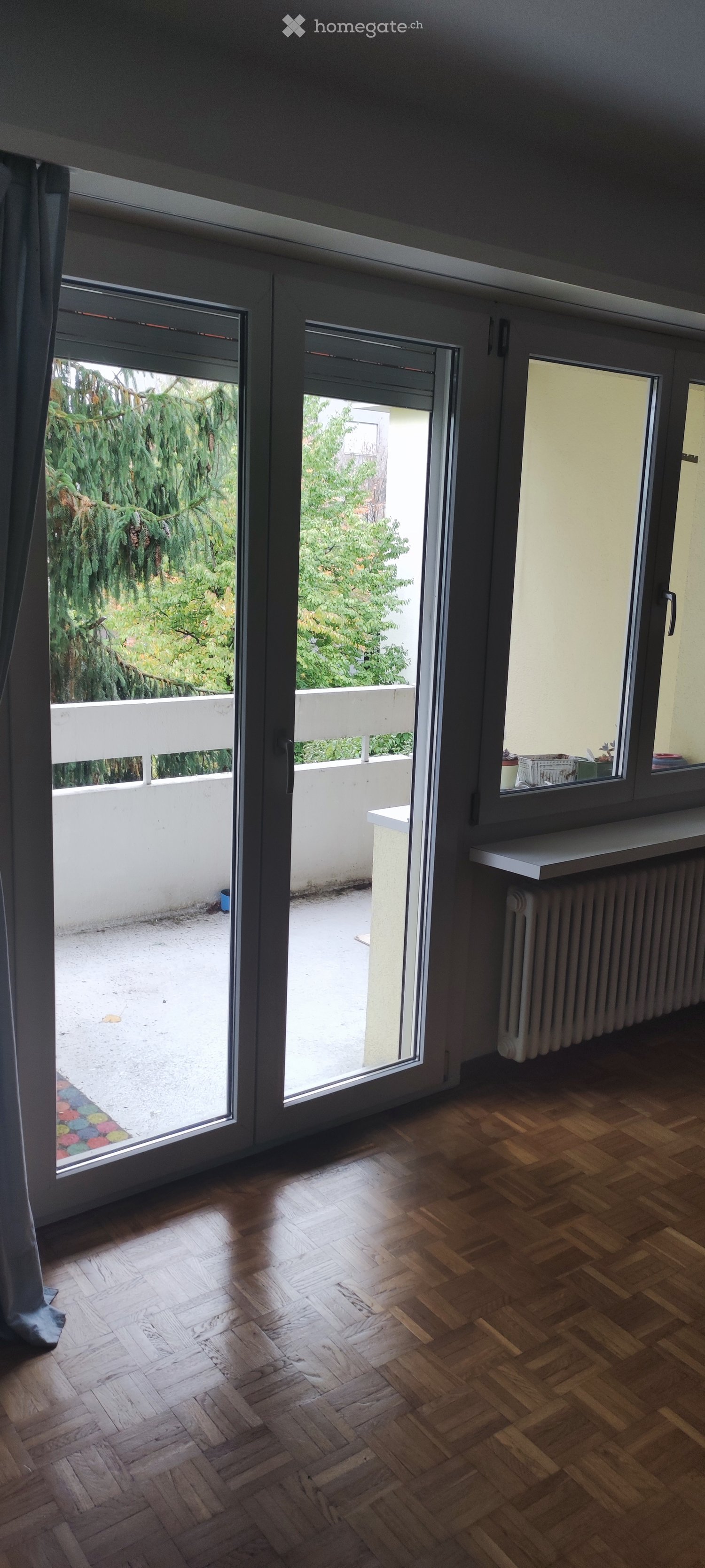 Empty room with wooden herringbone flooring, large sliding glass doors leading to a balcony, and a radiator on the wall.