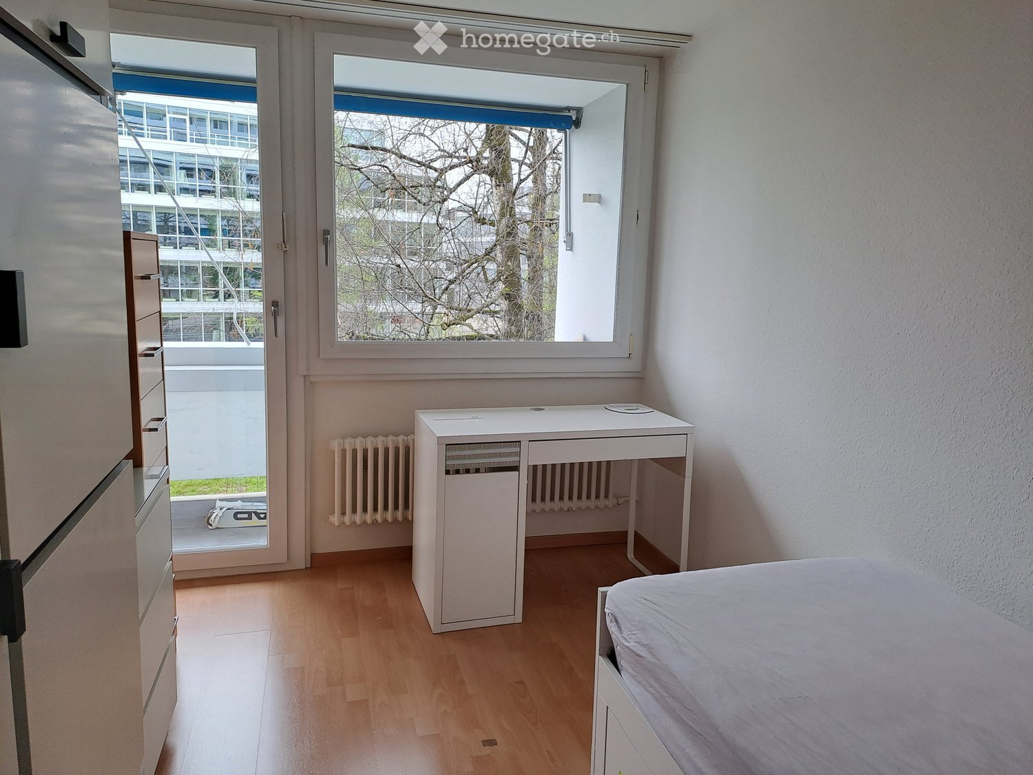 Modern, minimalist bedroom with light wooden flooring, white walls, a single bed, a small desk, and a radiator. Large window offers a view of the outside area.