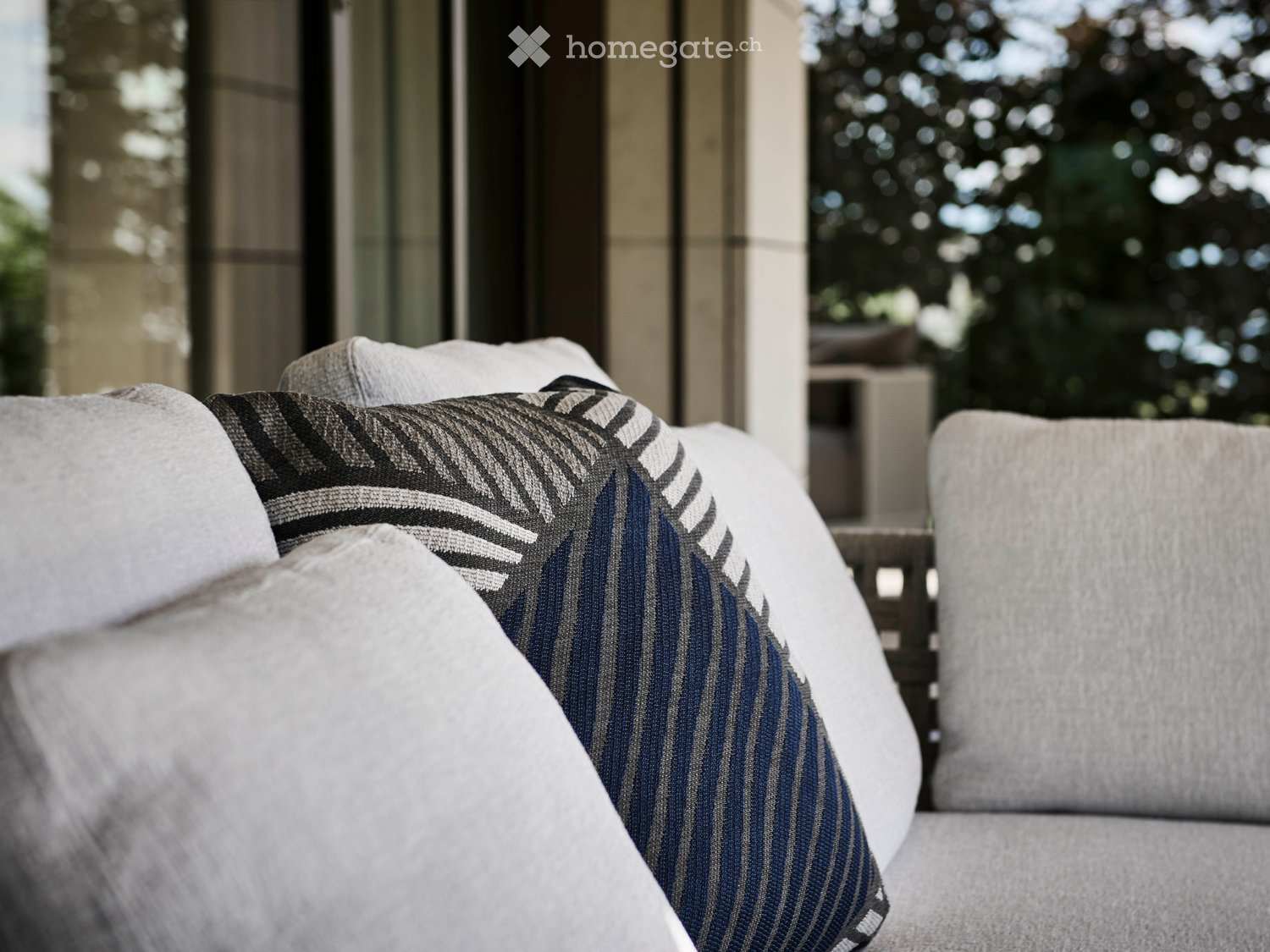 Modern outdoor seating area with grey sofa, patterned throw pillow, and white cushions, overlooking a lush garden.