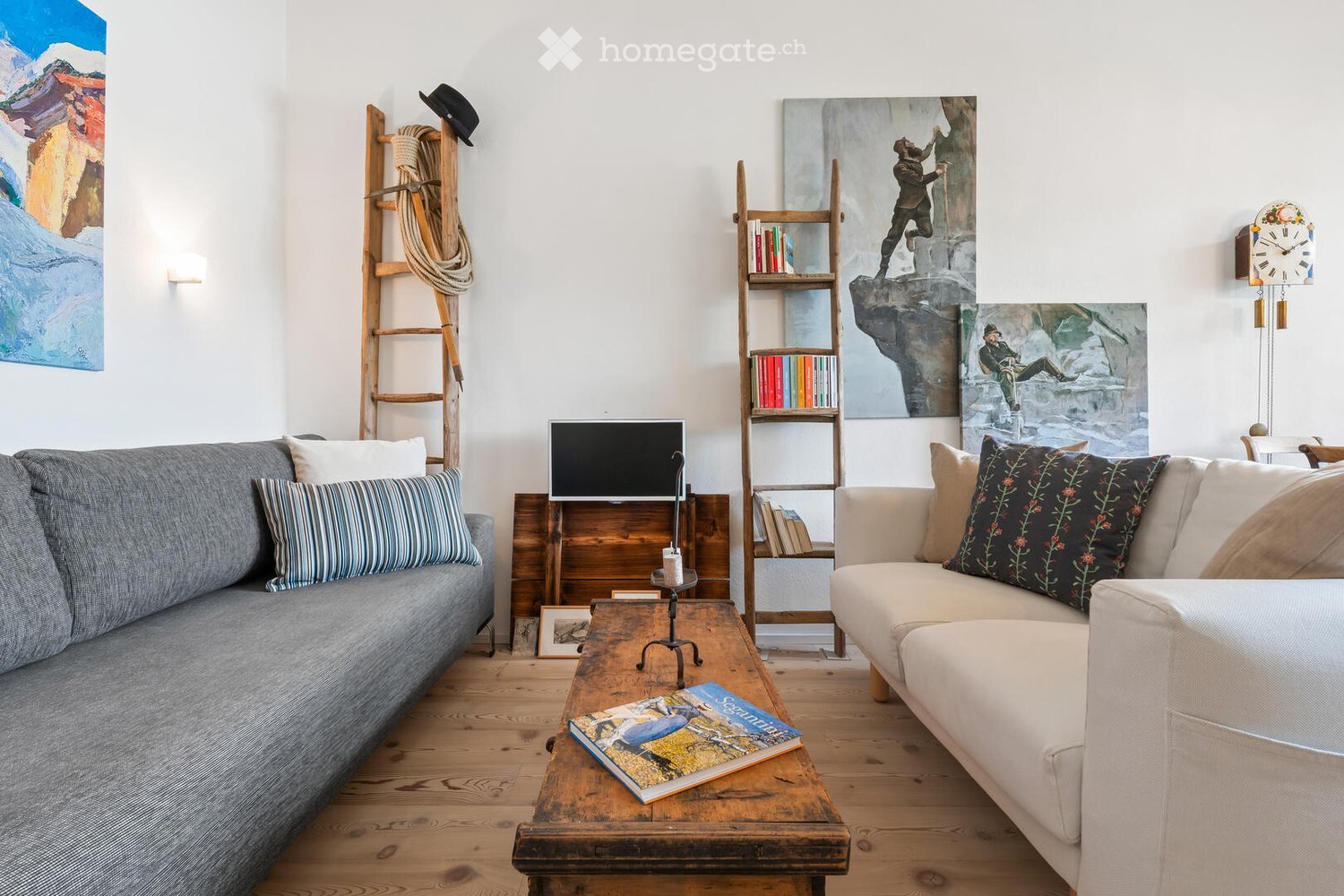 A cozy living room with wooden flooring, two sofas, a coffee table, a bookshelf, wall-mounted clock, and artwork on the walls.