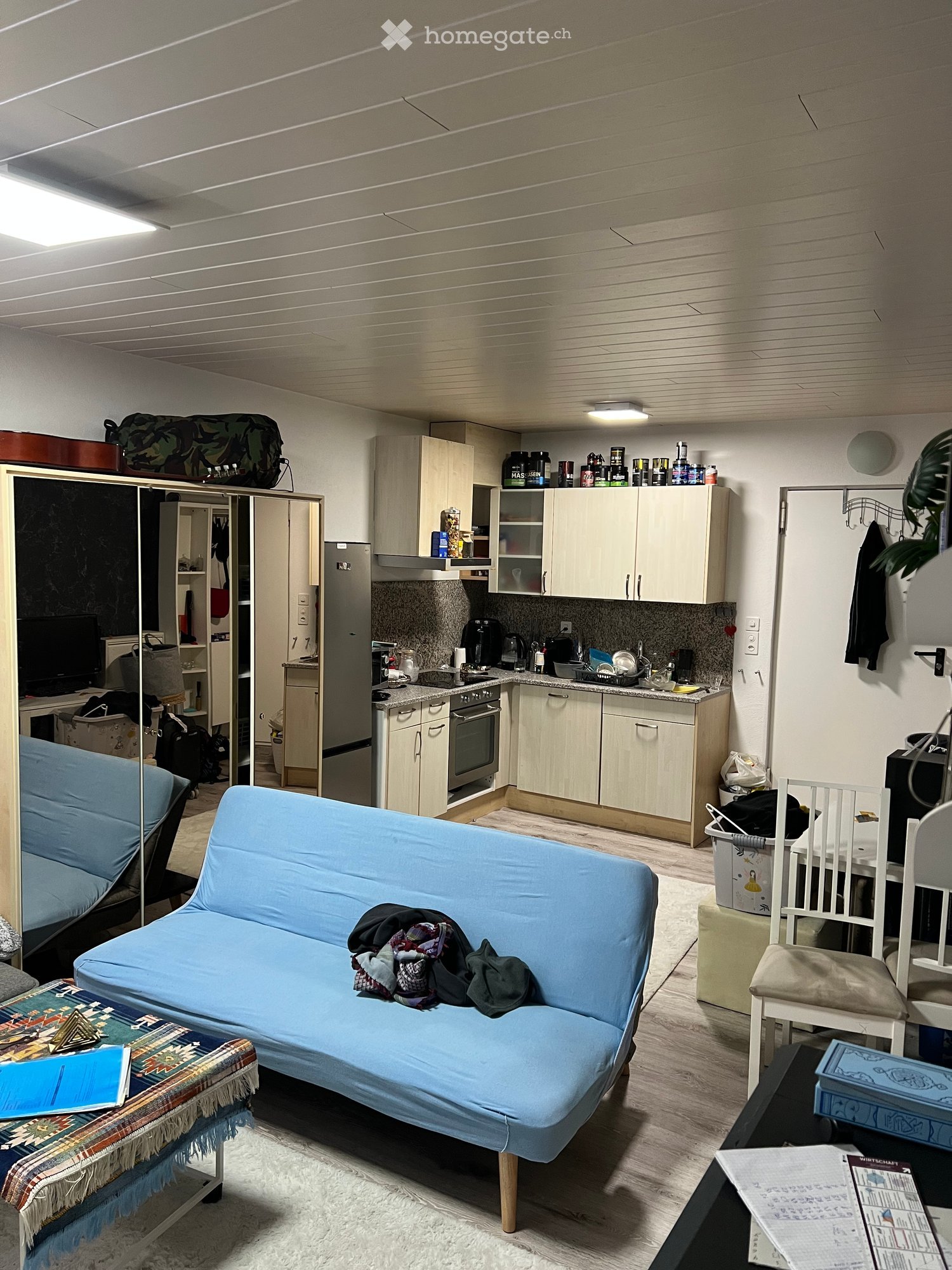 A modern, open-concept living and kitchen area with a blue sofa, a small table, a chair, and a kitchen equipped with cabinets, a refrigerator, an oven, and various appliances.