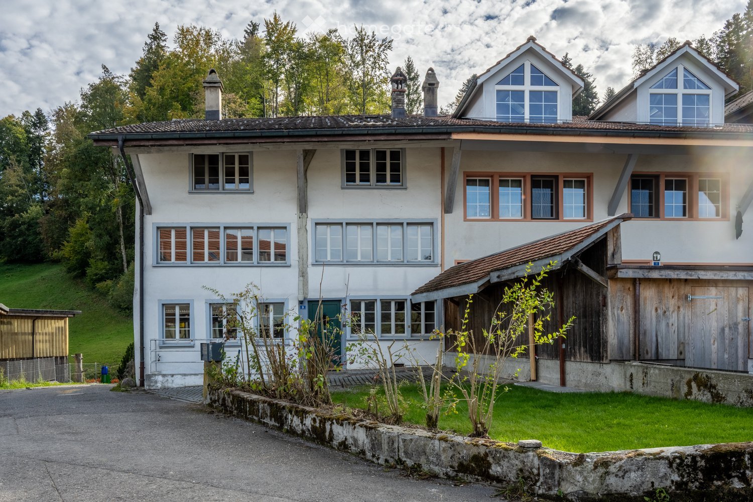 A two-story white house with multiple windows, a chimney, and a wooden garage. The house is surrounded by greenery and has a well-maintained lawn.