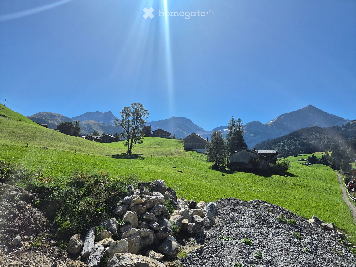 A picturesque alpine landscape with green hills, scattered houses, and distant mountains under a clear blue sky.