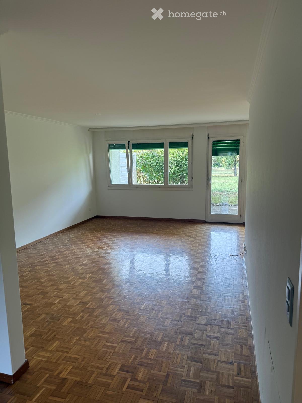 Empty room with wooden parquet flooring, white walls, large windows, and a sliding glass door leading to an outdoor area.