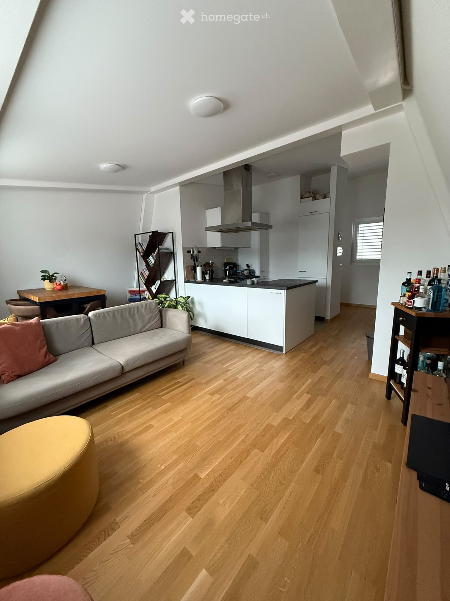 Modern living room with light wooden flooring, grey sofa, round yellow stool, small wooden table, bookshelf, and an open kitchen area with white cabinets and stainless steel appliances.