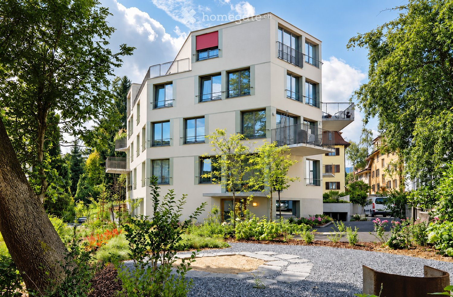 Modern multi-story residential building with balconies, surrounded by greenery and a landscaped garden area.