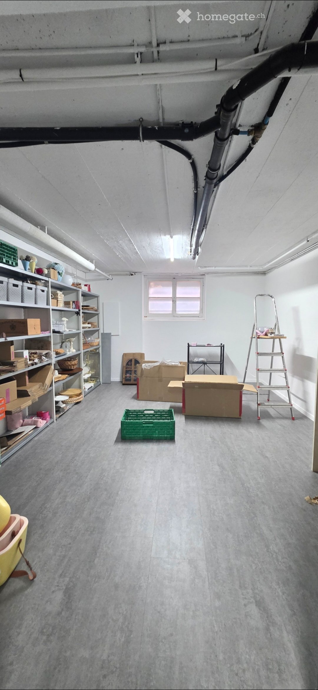 An attic or storage room with exposed pipes and beams, featuring a large window, shelves filled with various items, and several cardboard boxes on the floor.
