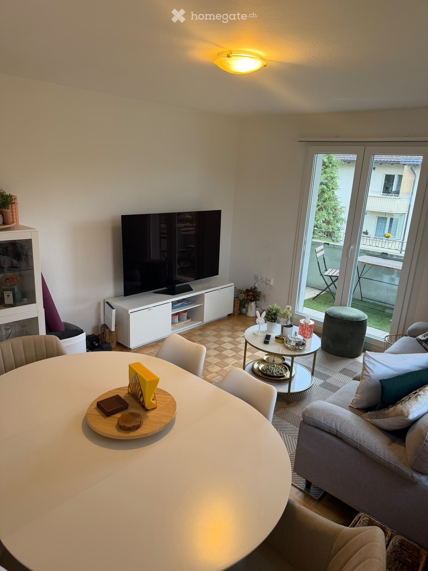 Modern living room with a grey sofa, coffee table, round dining table, chairs, and a television. Features a balcony with outdoor seating visible through large glass doors.