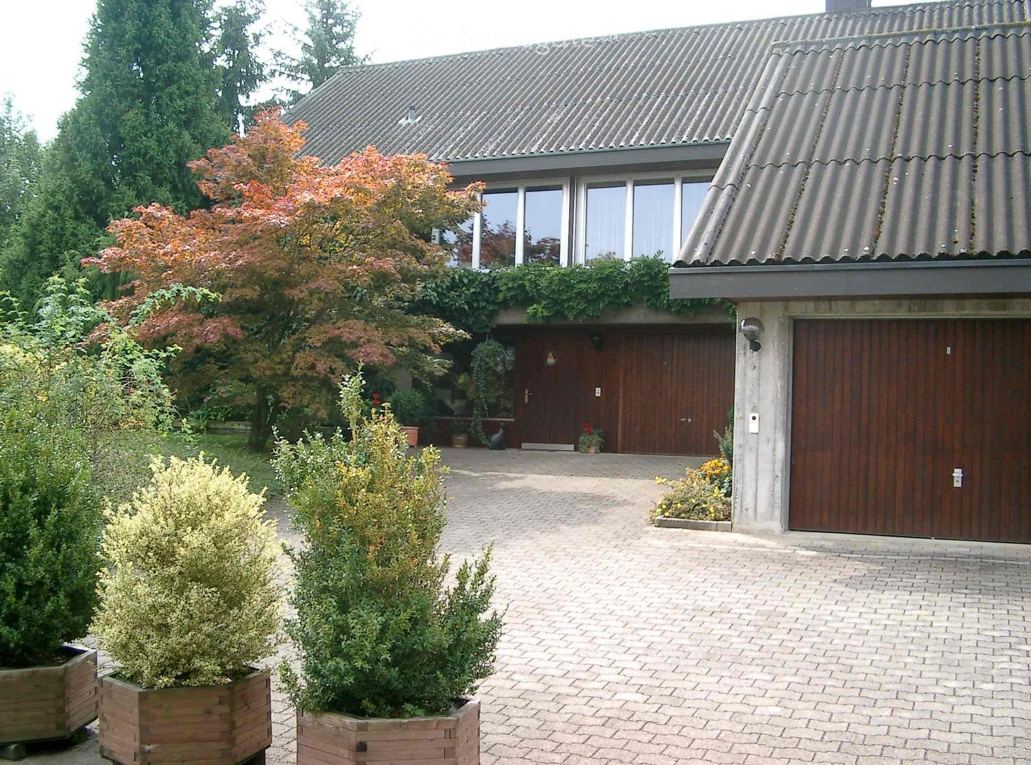 Two-story house with a grey tiled roof, large windows, and a wooden garage door. The entrance features a wooden door flanked by potted plants and shrubs, with a well-maintained garden and driveway.
