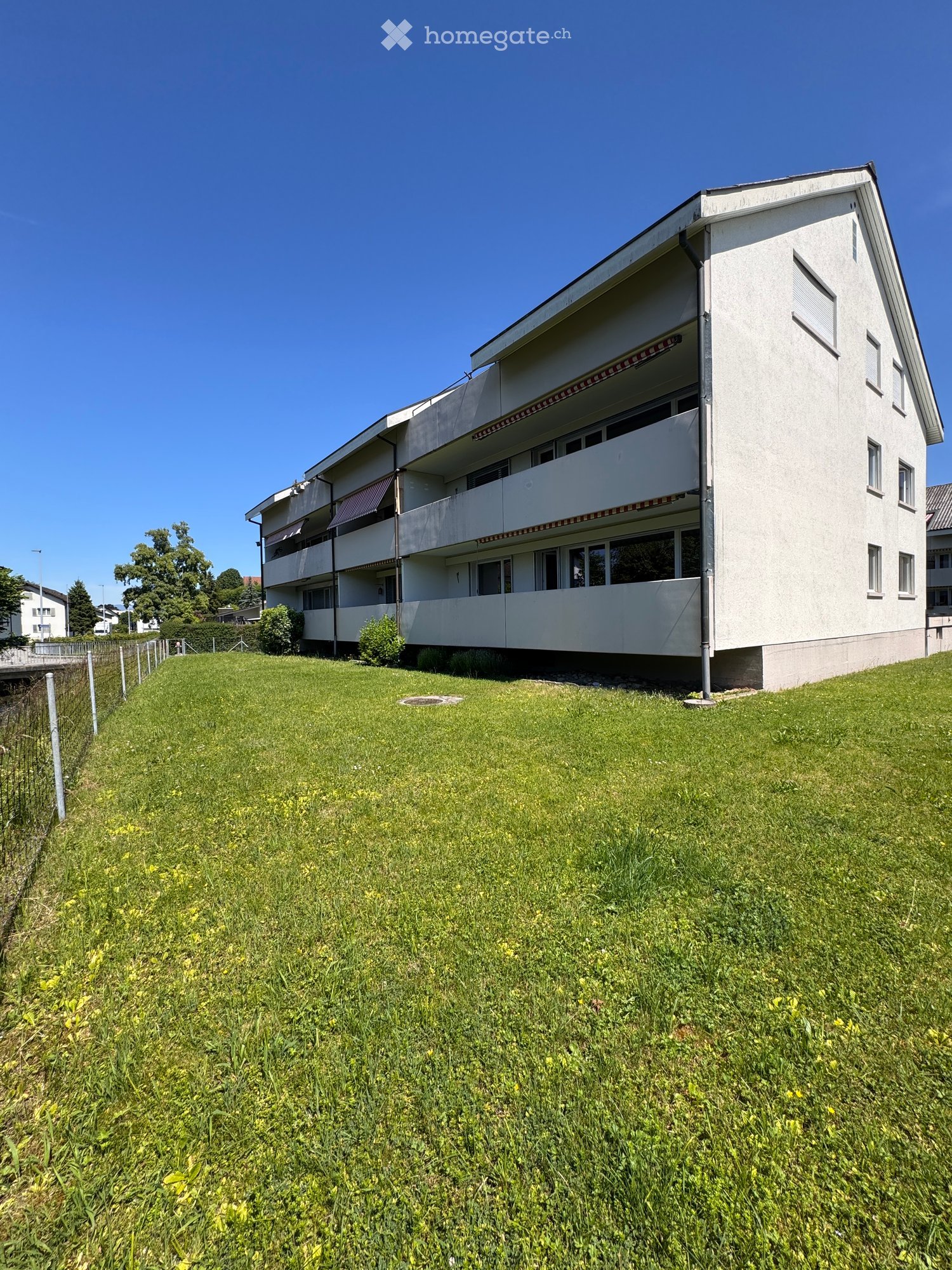 A modern multi-story apartment building with white walls, large windows, and balconies, situated on a grassy slope.