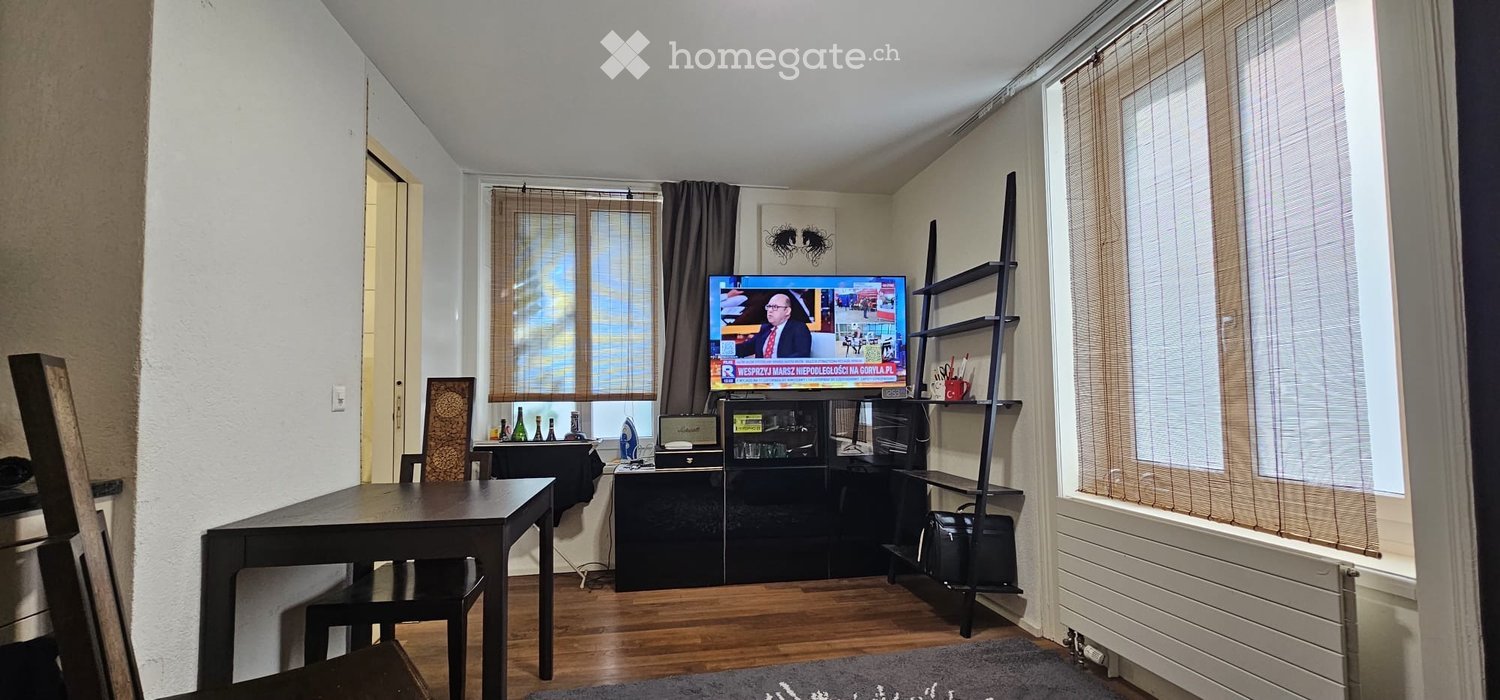 A modern living room with a wooden floor, large windows with blinds, a TV on a stand, a dining table with chairs, and a shelf with decorative items.