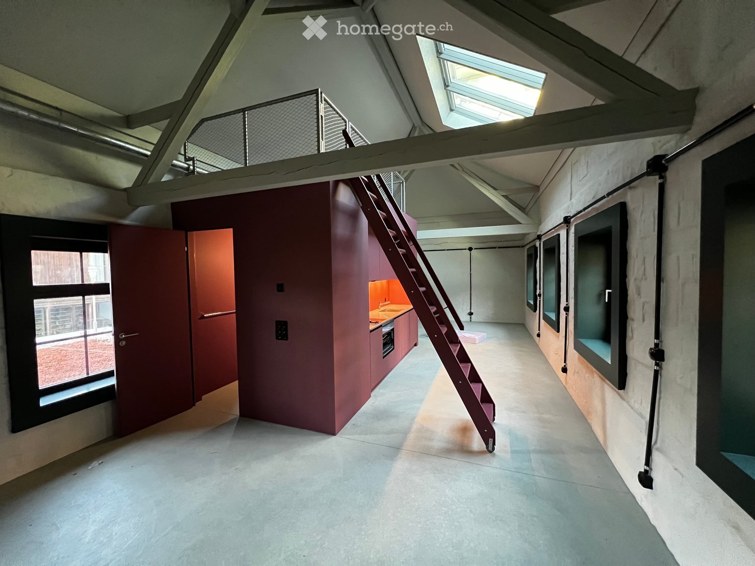 An empty, modern loft-style room with exposed wooden beams, a metal staircase leading to an upper level, and a small kitchen area with cabinets and a sink.