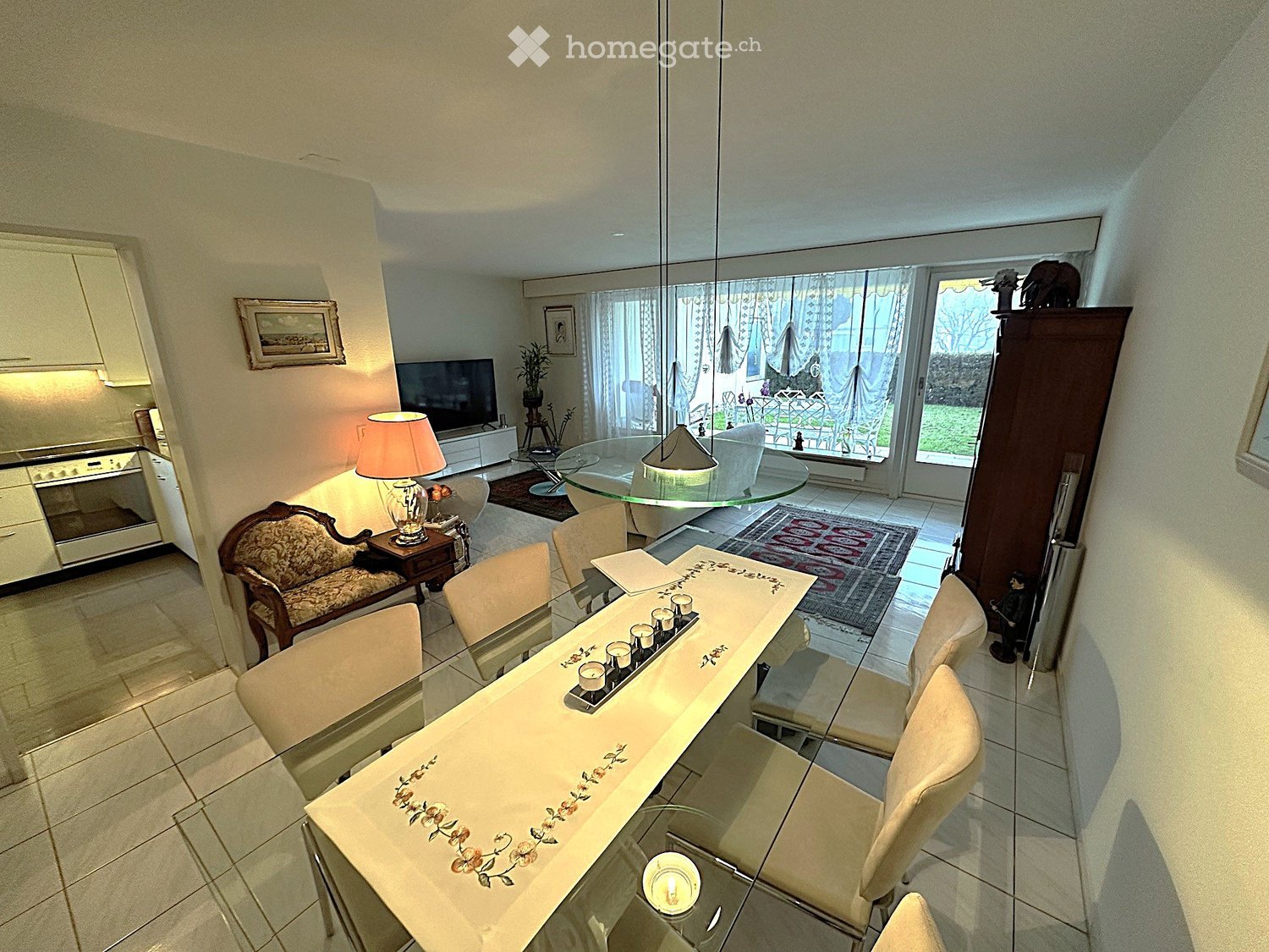 A well-lit living and dining area with a glass dining table, chairs, a lamp, a sofa, a television, and an oven in the background.