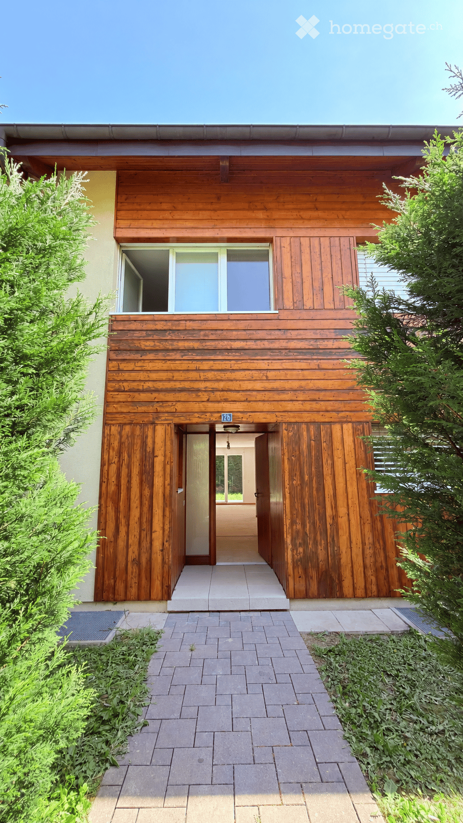 Modern two-story house with wooden exterior, large windows, and a covered entrance area. The house features a raised ground floor and is surrounded by greenery and a paved pathway.