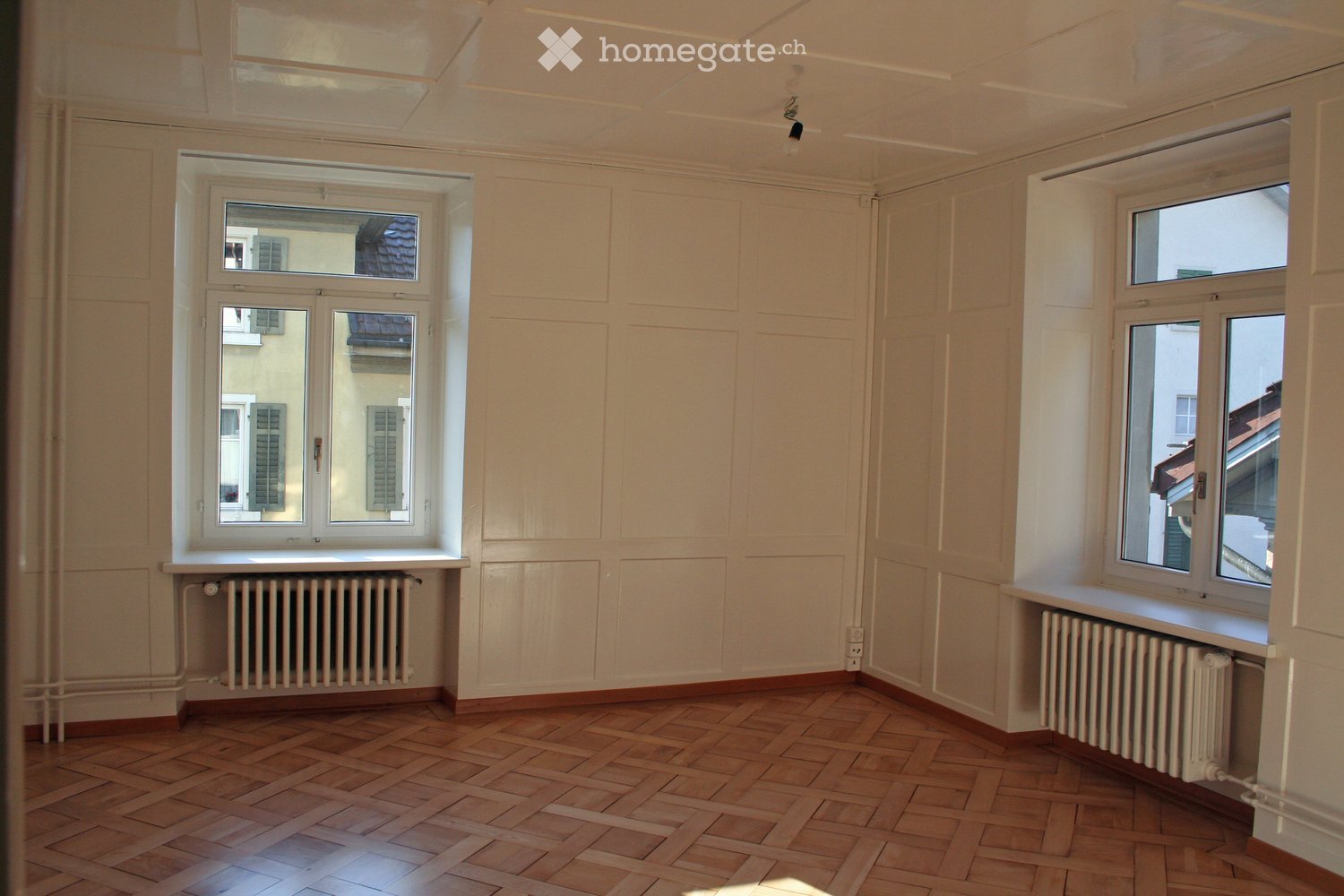 Empty room with wooden parquet flooring, white paneled walls, and large windows with radiators beneath them.