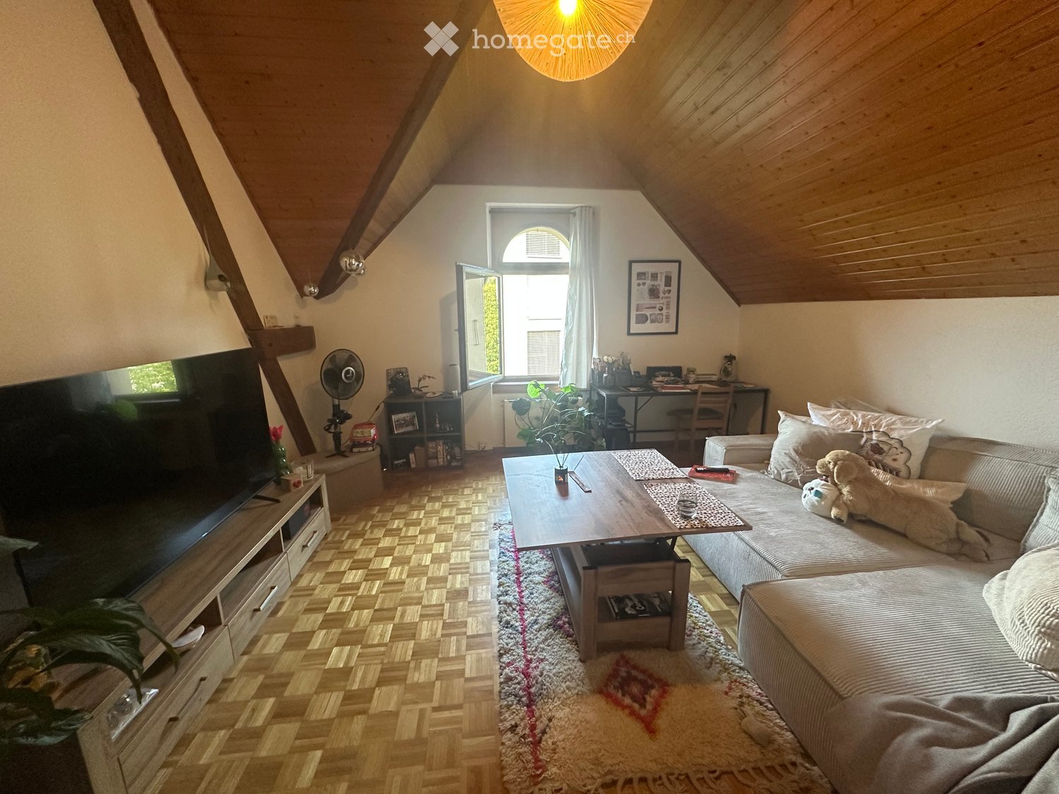 Cozy living room with slanted wooden ceiling, large window, sofa, coffee table, TV, and small desk area.