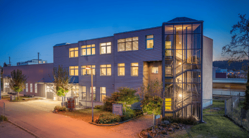 Modern multi-story building with illuminated windows and a glass staircase, surrounded by landscaping and a paved driveway.