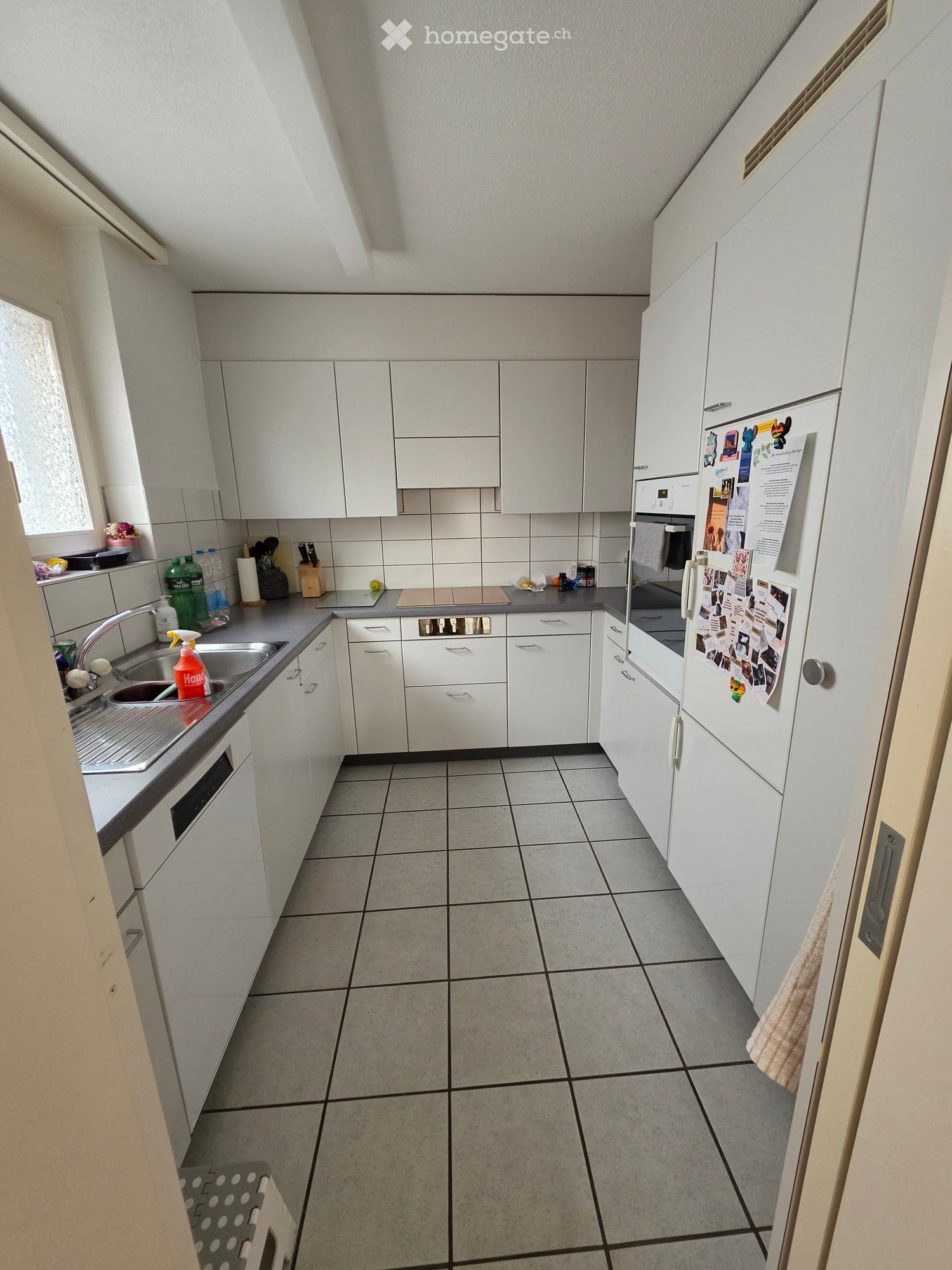 Modern kitchen with white cabinets, tiled floor, stainless steel sink, refrigerator, and countertop items including dish soap and cutting board.