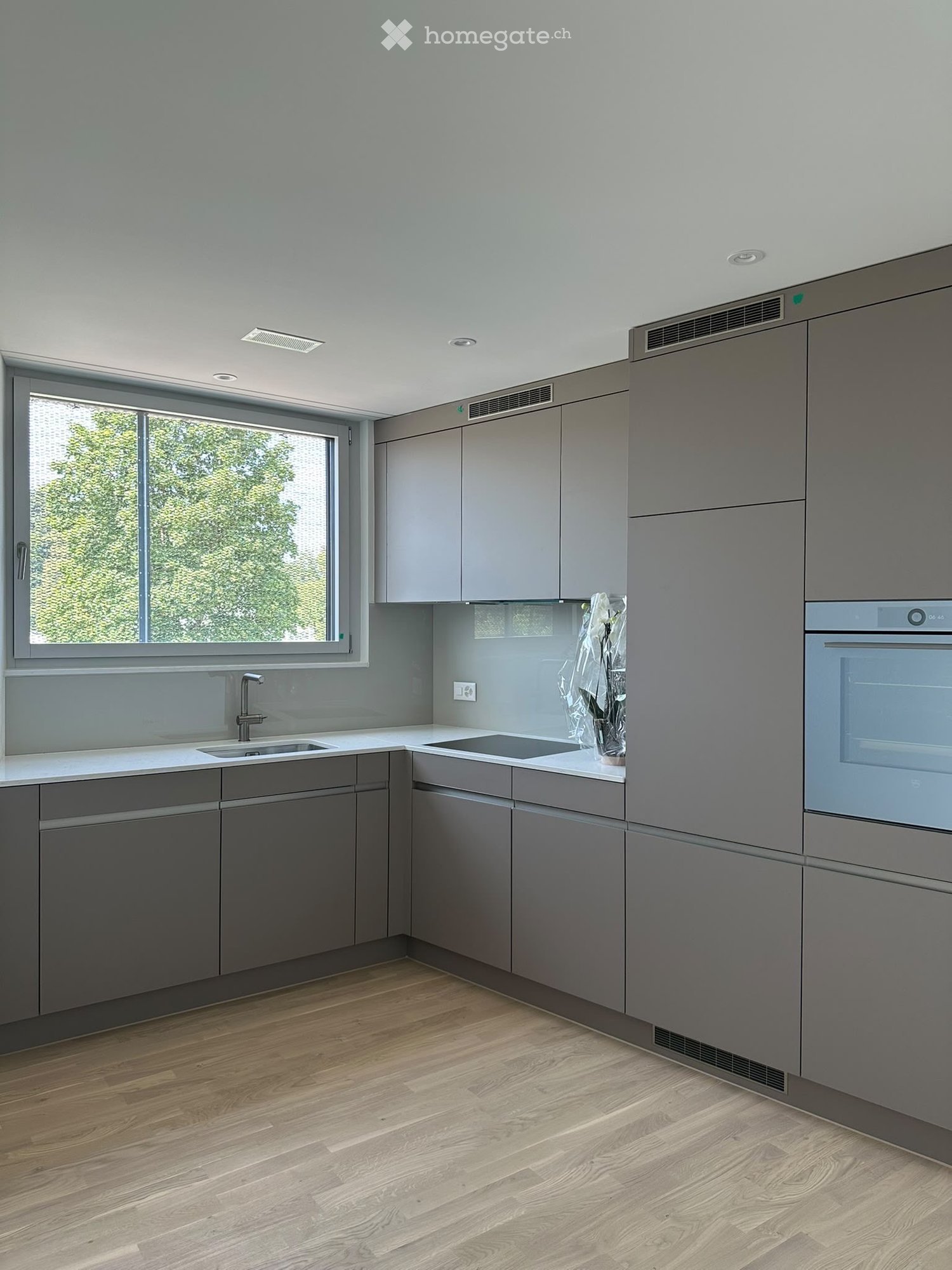 Modern kitchen with grey cabinets, white countertops, wooden flooring, and a large window overlooking greenery.