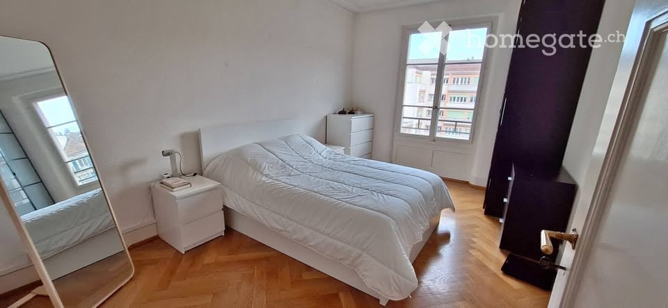 Bright bedroom with wooden flooring, large window offering a city view, a neatly made bed, a nightstand with a lamp, and a full-length mirror.