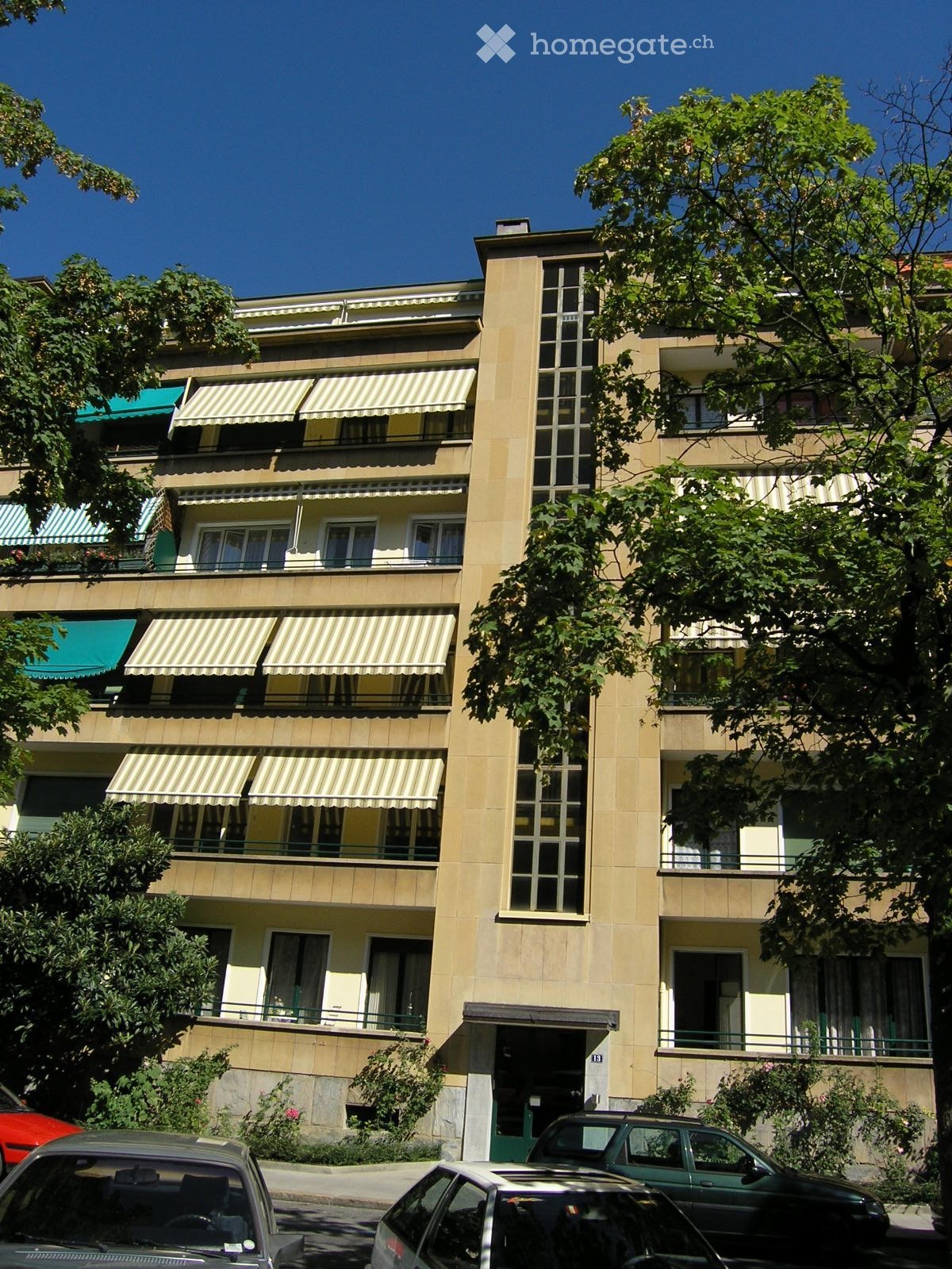 Multi-story residential building with beige facade, balconies, striped awnings, and surrounding greenery.