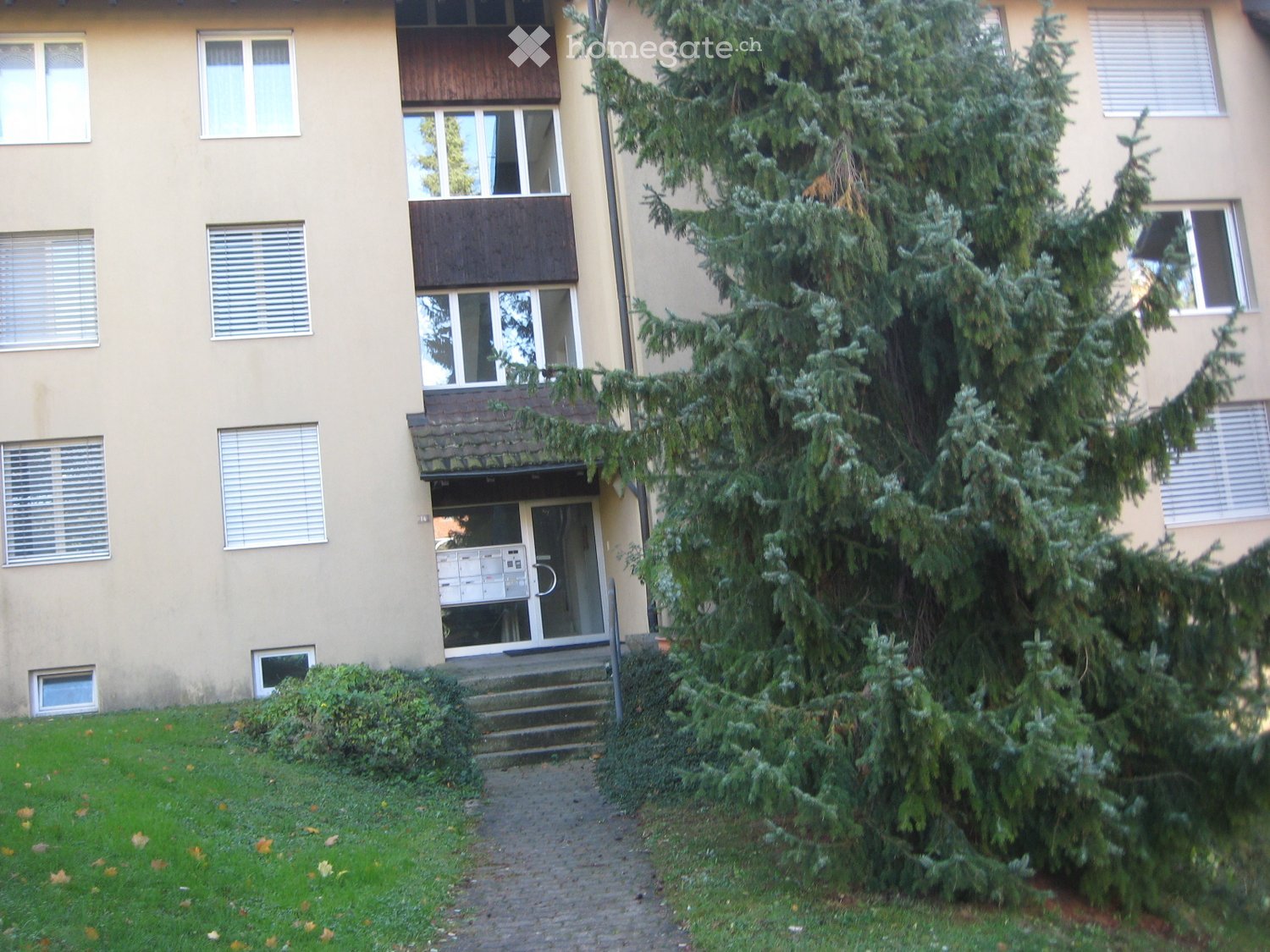 Multi-story residential building with beige facade, multiple windows, balconies, and a small garden area with trees and shrubs.