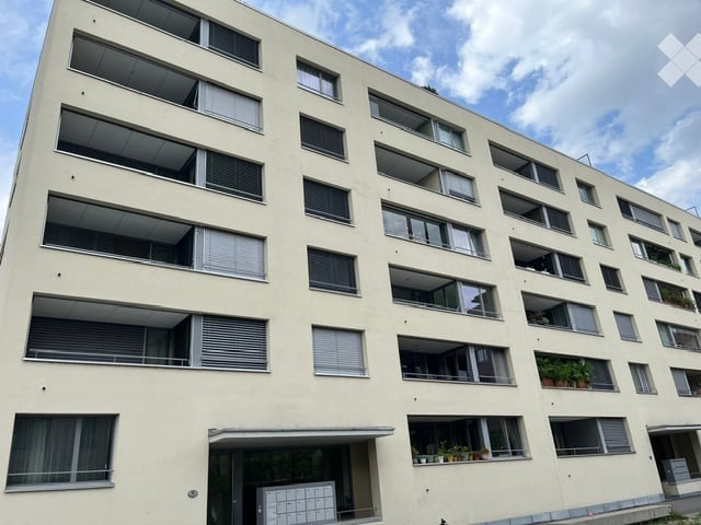 Modern multi-story residential building with numerous windows and balconies, light-colored facade, and a small entrance area at the bottom.