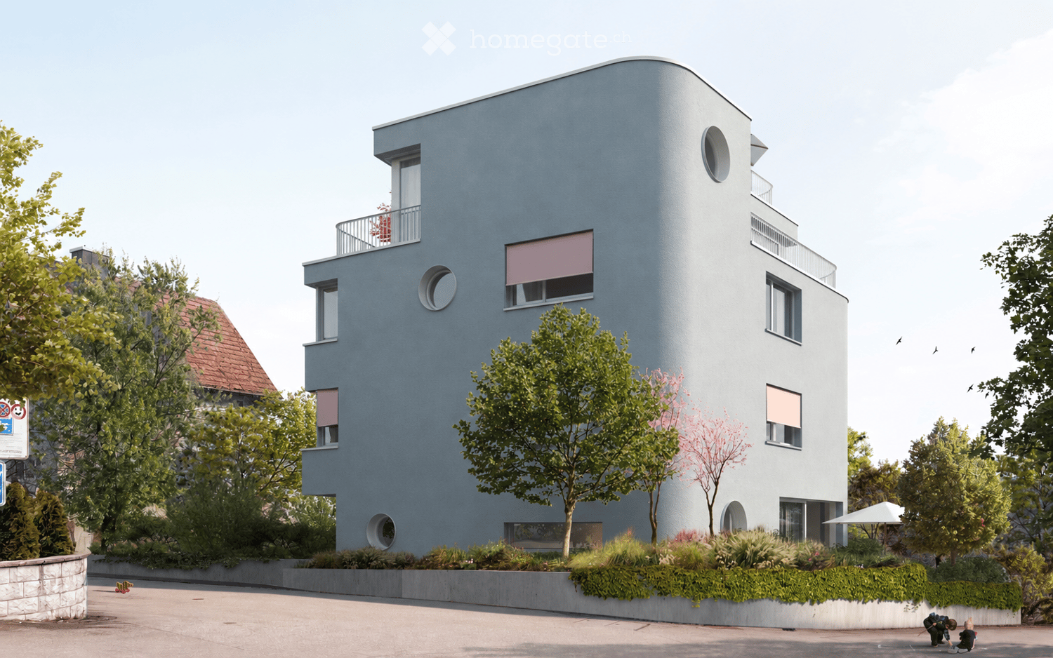 Modern, two-story building with a sleek, curved facade in light blue, featuring large windows and balconies. The building is surrounded by greenery and trees, with a raised ground floor and a small garden area in front.