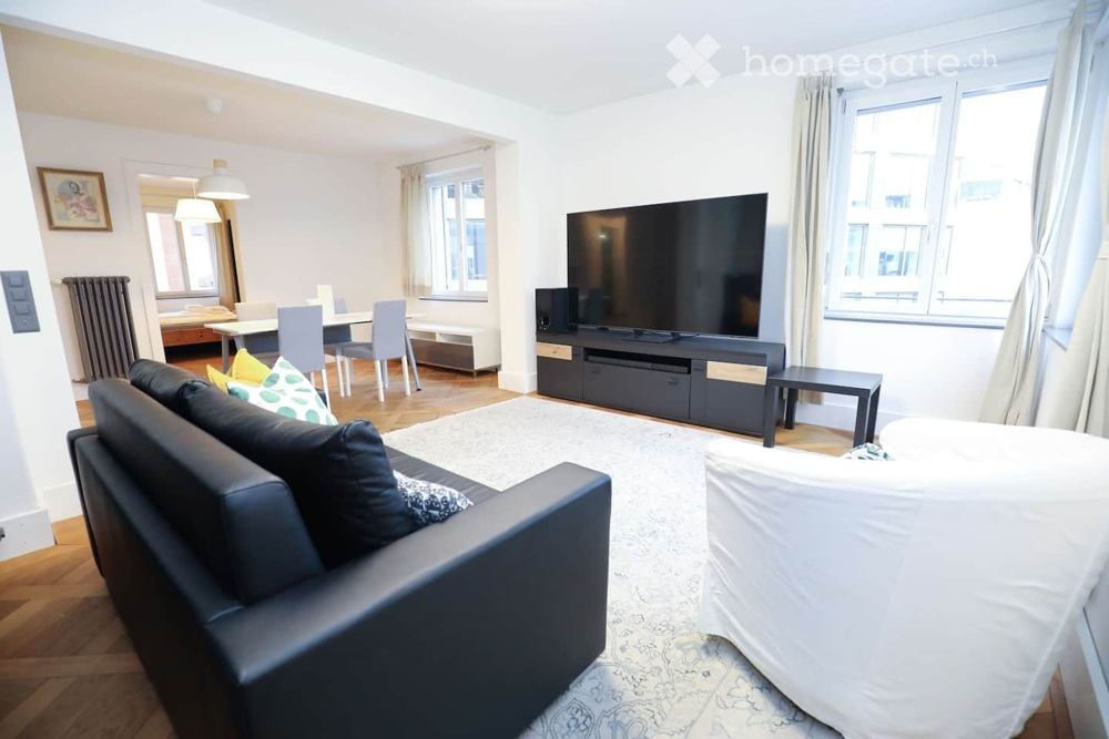 Modern living room with a black leather sofa, ottoman, coffee table, TV, dining area with table and chairs, and large windows with curtains.