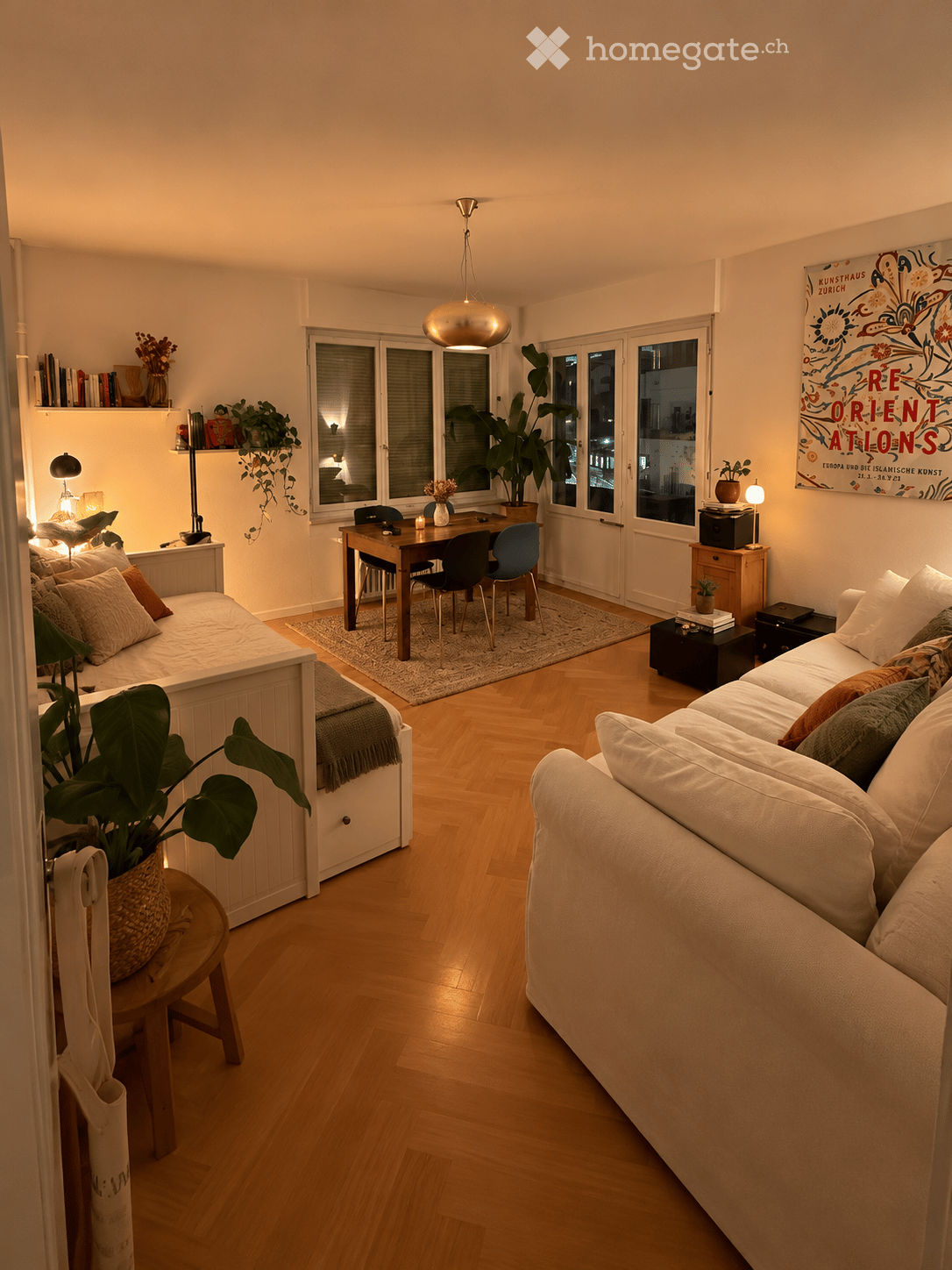 A modern, well-lit living room with a white sofa, wooden dining table, chairs, and a hanging pendant light. The room features wooden flooring, large windows, and decorative plants.